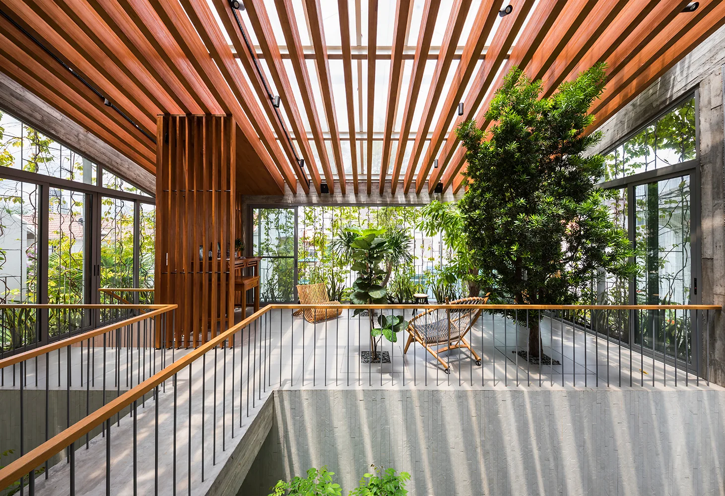 An indoor-outdoor terrace featuring a slatted wooden ceiling, large trees planted directly in the tiled floor, and floor-to-ceiling glass walls.