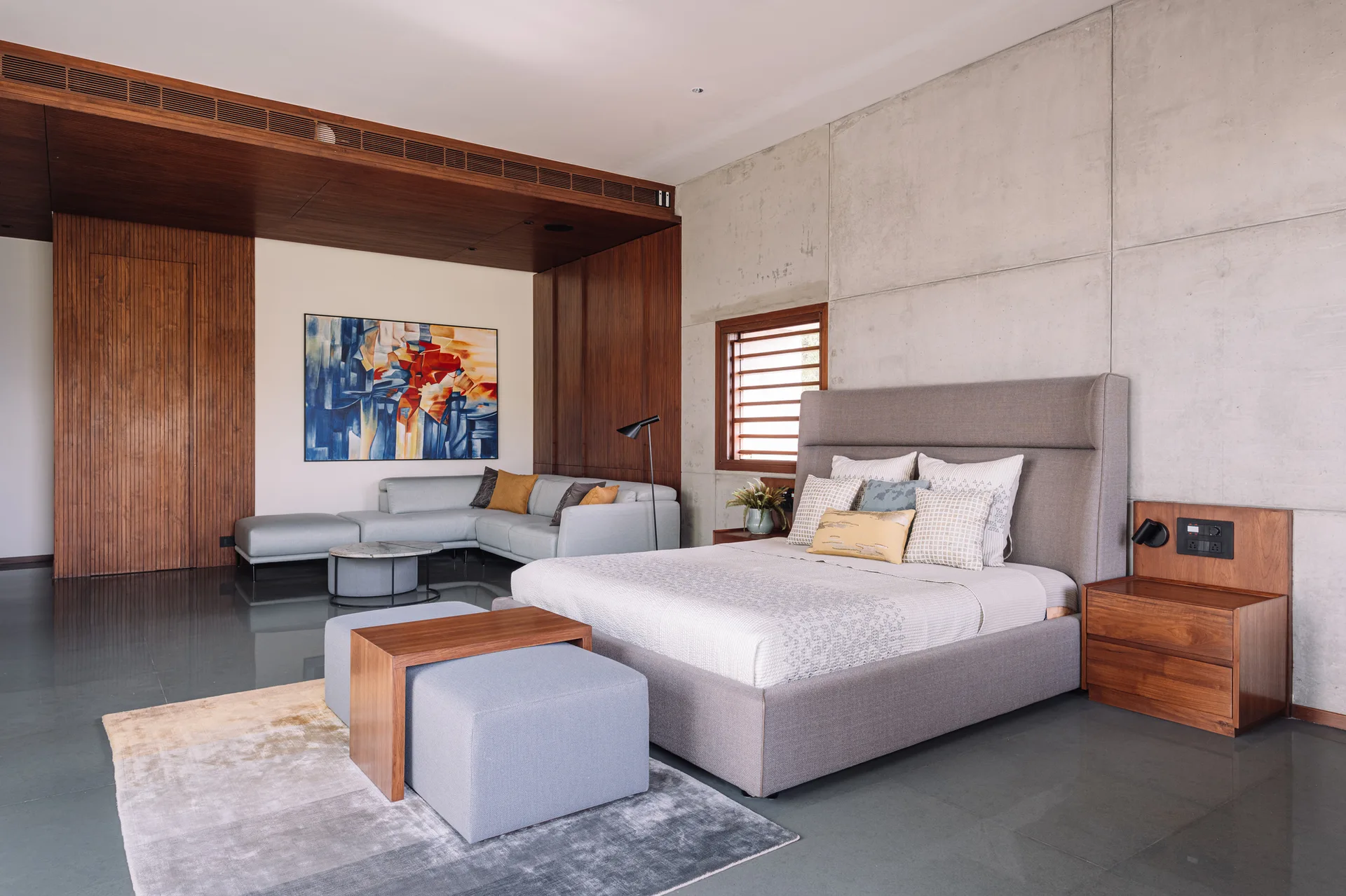 A modern bedroom featuring a grey upholstered bed against a concrete panel wall next to a wood-paneled alcove with a sectional sofa.