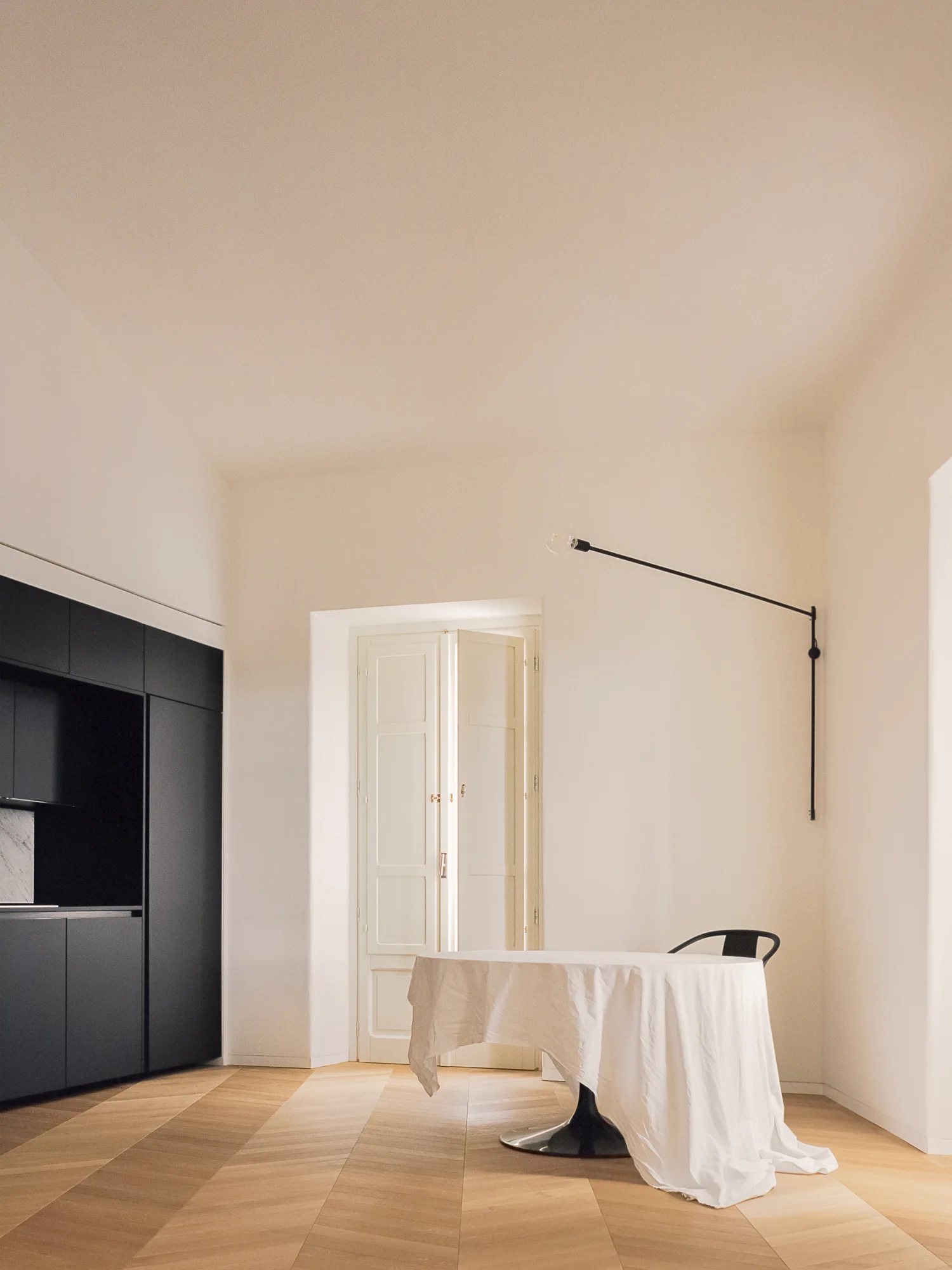 A minimalist dining room features a round table draped in a white cloth, a black wall lamp, matte black cabinetry, and chevron wood flooring.