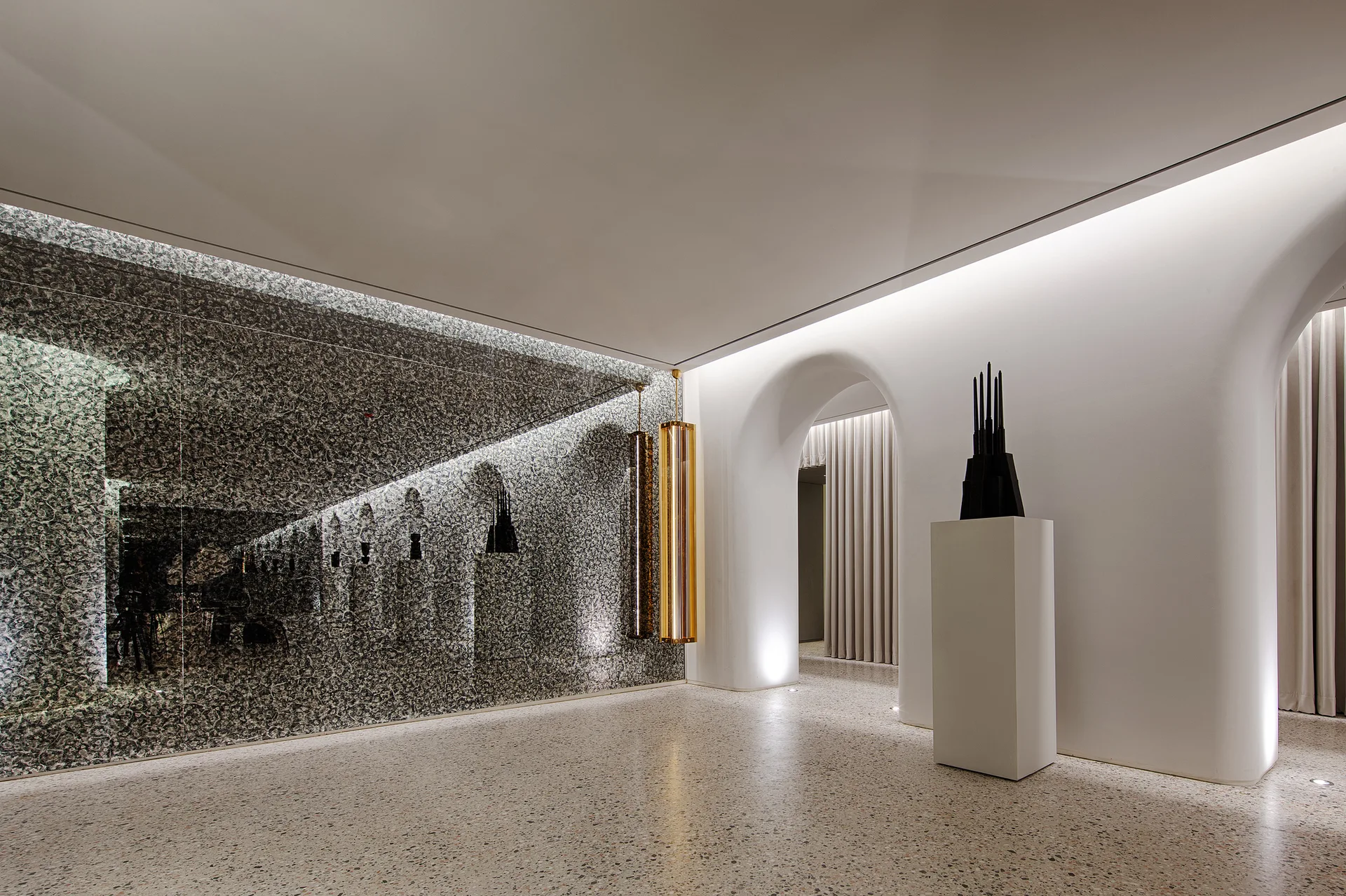 A minimalist hallway with white arched doorways, polished terrazzo flooring, a dark patterned feature wall, and a black sculpture on a rectangular pedestal.