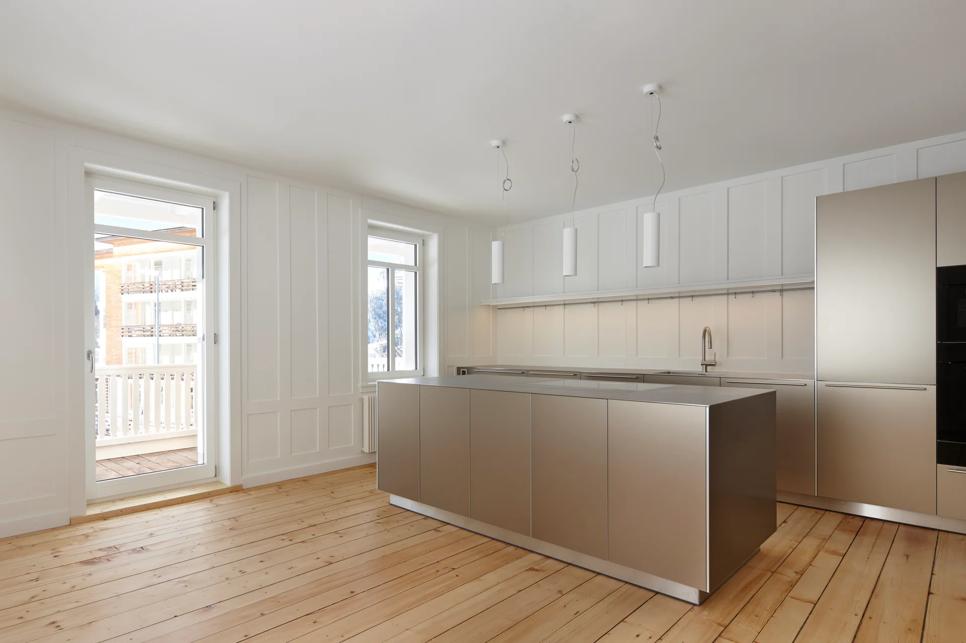 A minimalist kitchen features a metallic island and cabinetry, white paneled walls, light wood plank floors, and three white pendant lights.