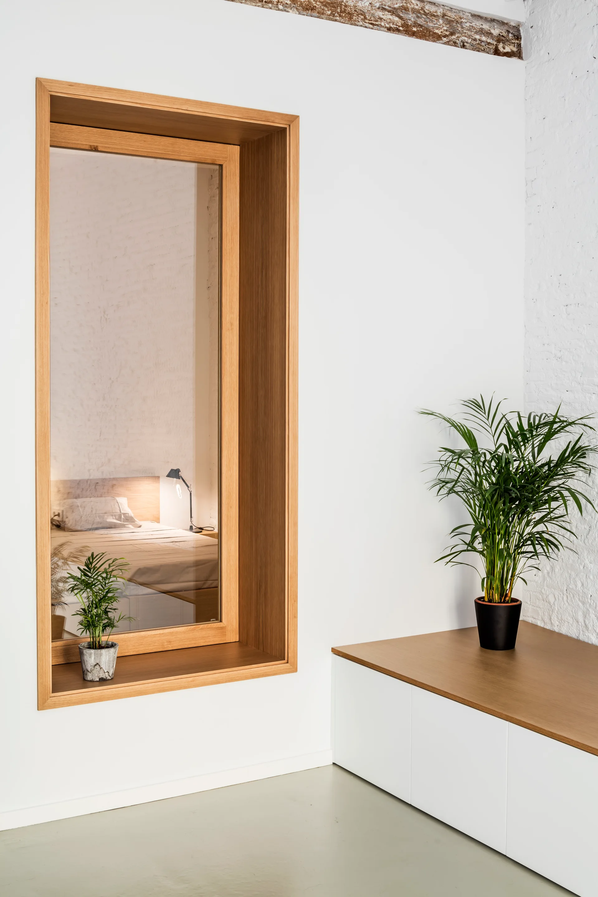 A large wood-framed internal window looks into a bedroom next to a white built-in cabinet with a potted palm.