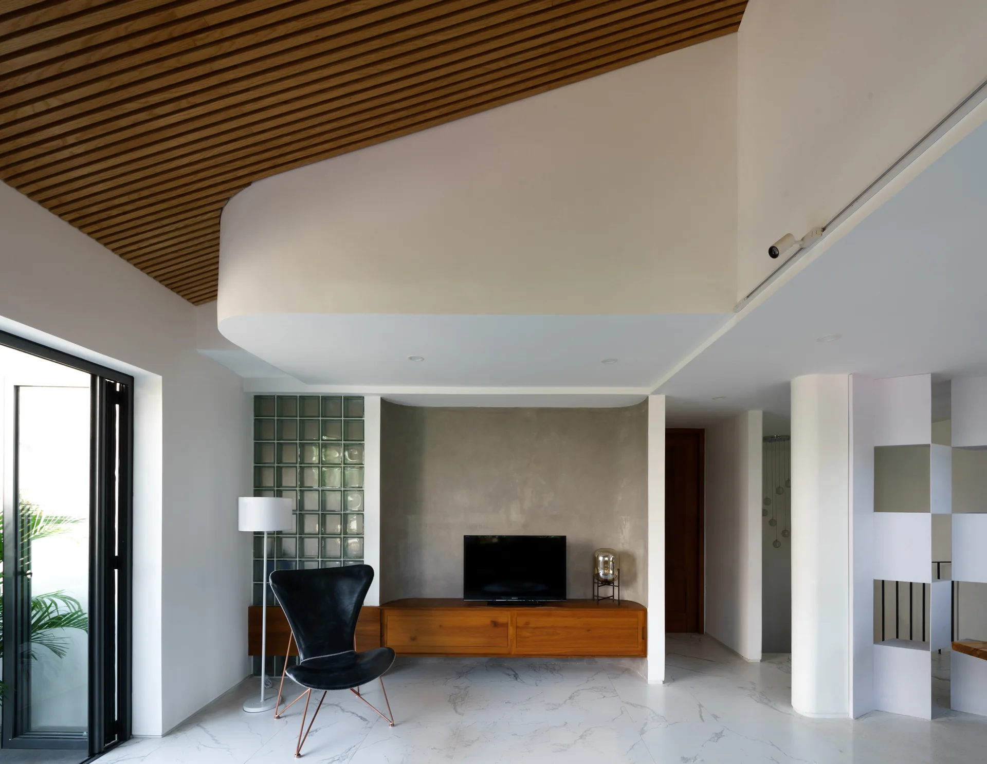 A modern living room features a sloped wooden slat ceiling, a glass block partition wall, a low wood media console, and a black leather chair.