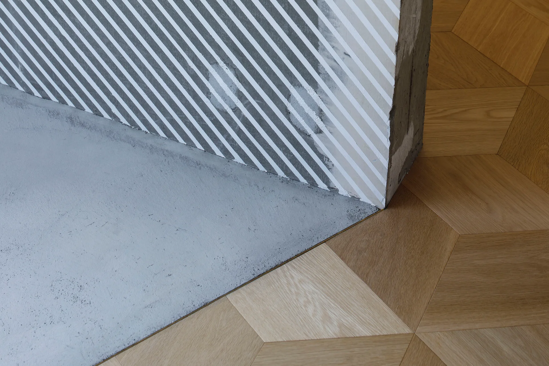 A close-up of a floor transition where geometric light oak parquet meets a smooth grey concrete surface next to a striped wall.