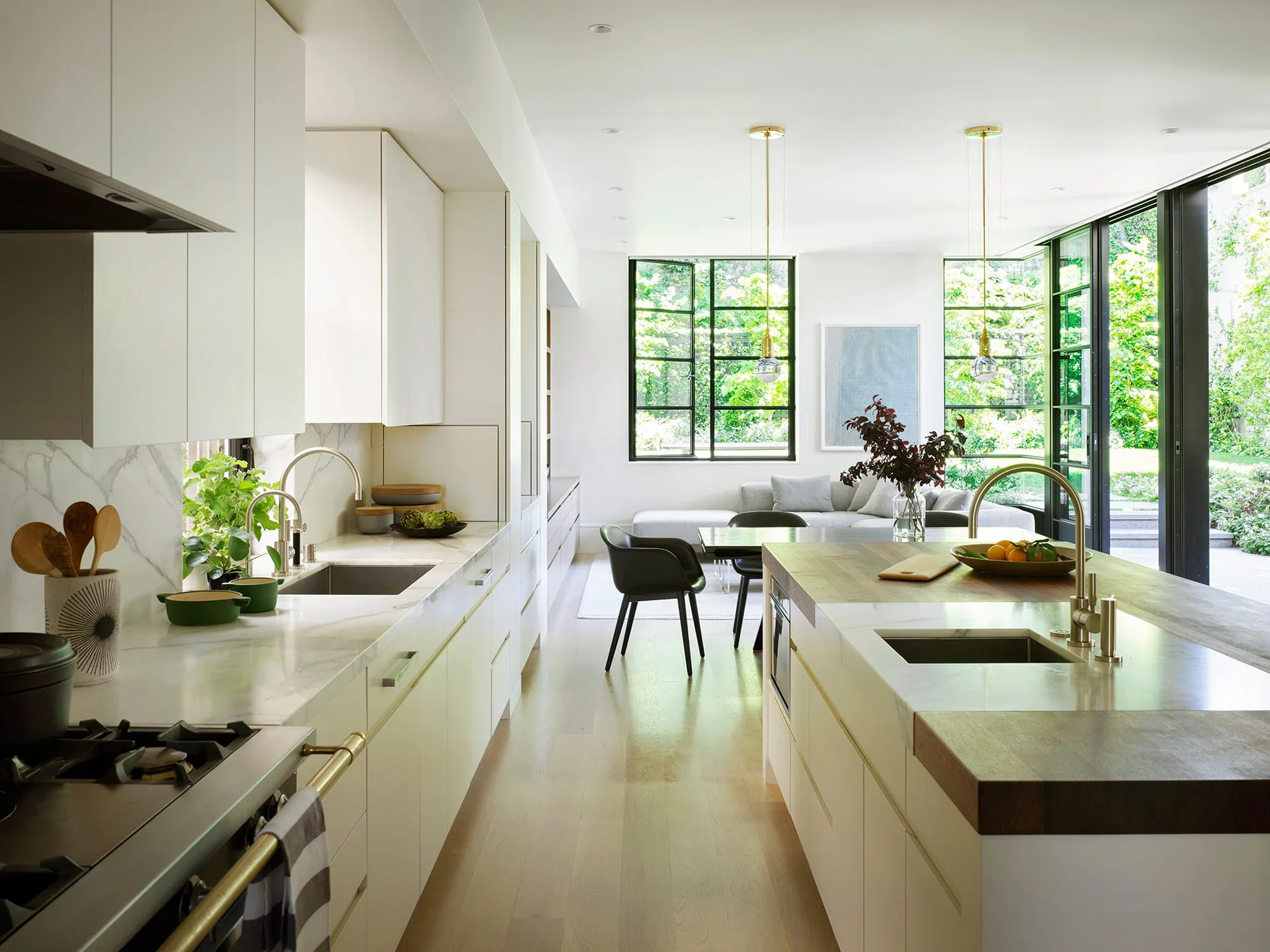 A modern open-concept kitchen features white flat-front cabinets, a marble and wood island, and large black-framed windows overlooking a green garden.