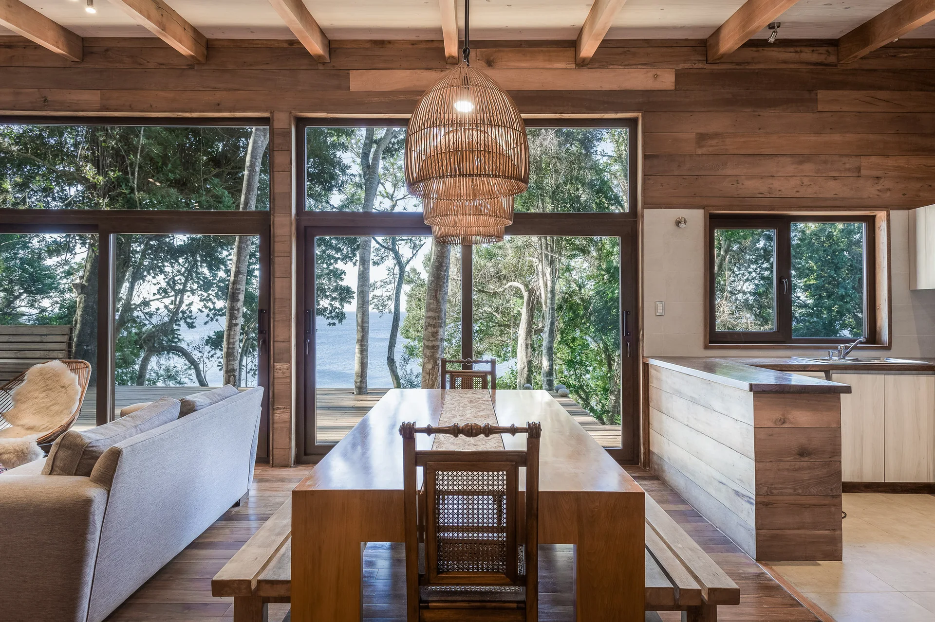 A rustic wood-paneled dining room features a long wooden table, a large woven rattan pendant light, and floor-to-ceiling windows overlooking trees and water.