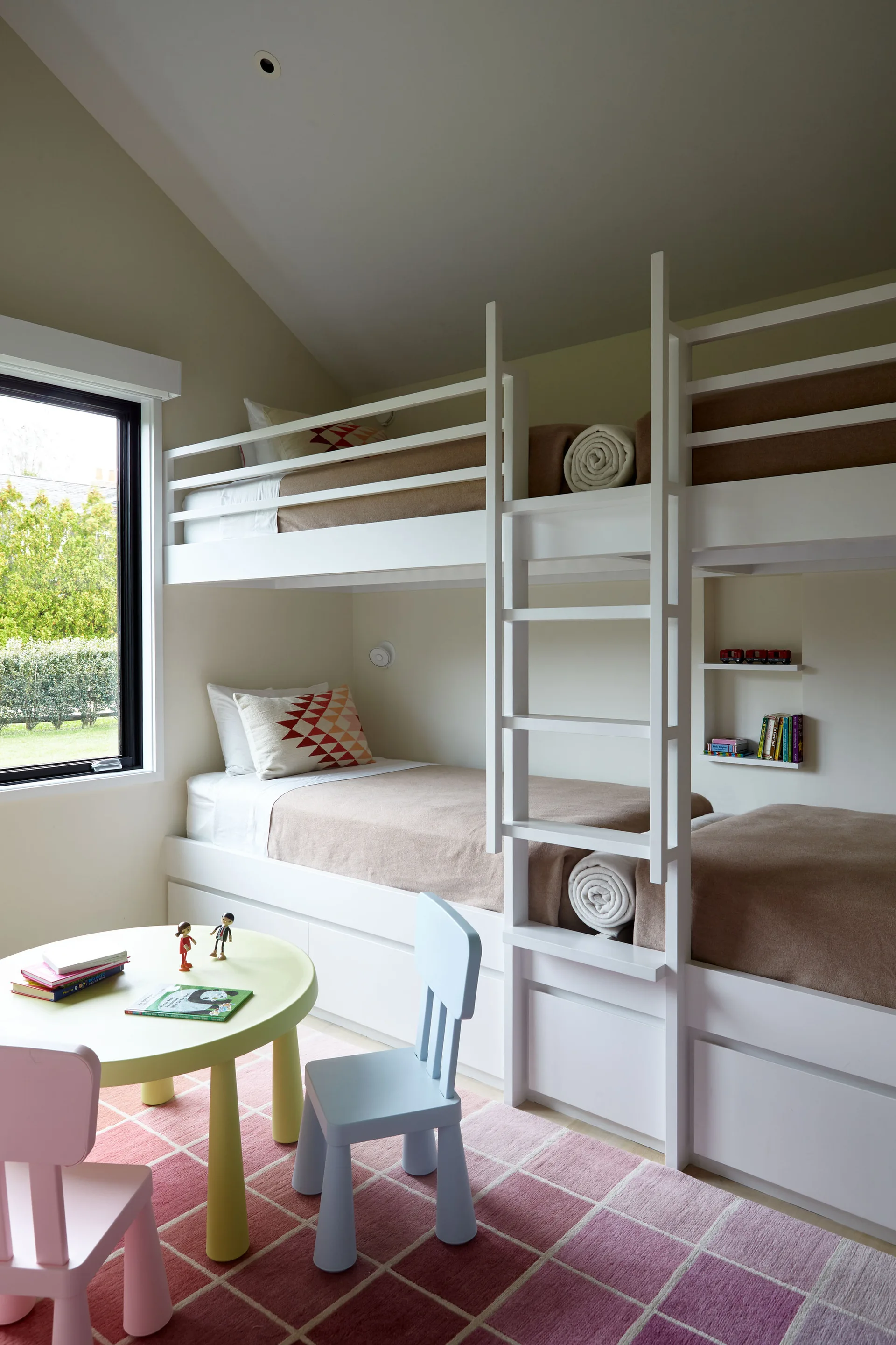 A children's bedroom features a white built-in bunk bed, a small round yellow table with pastel chairs, and a pink grid rug under a sloped ceiling.