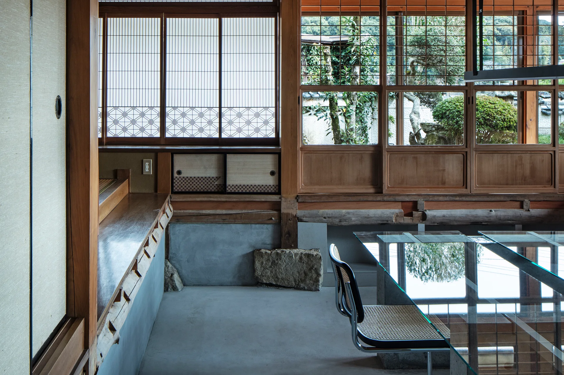 A minimalist Japanese interior with wooden shoji windows, a smooth concrete floor, and a modern glass table with a cane chair.