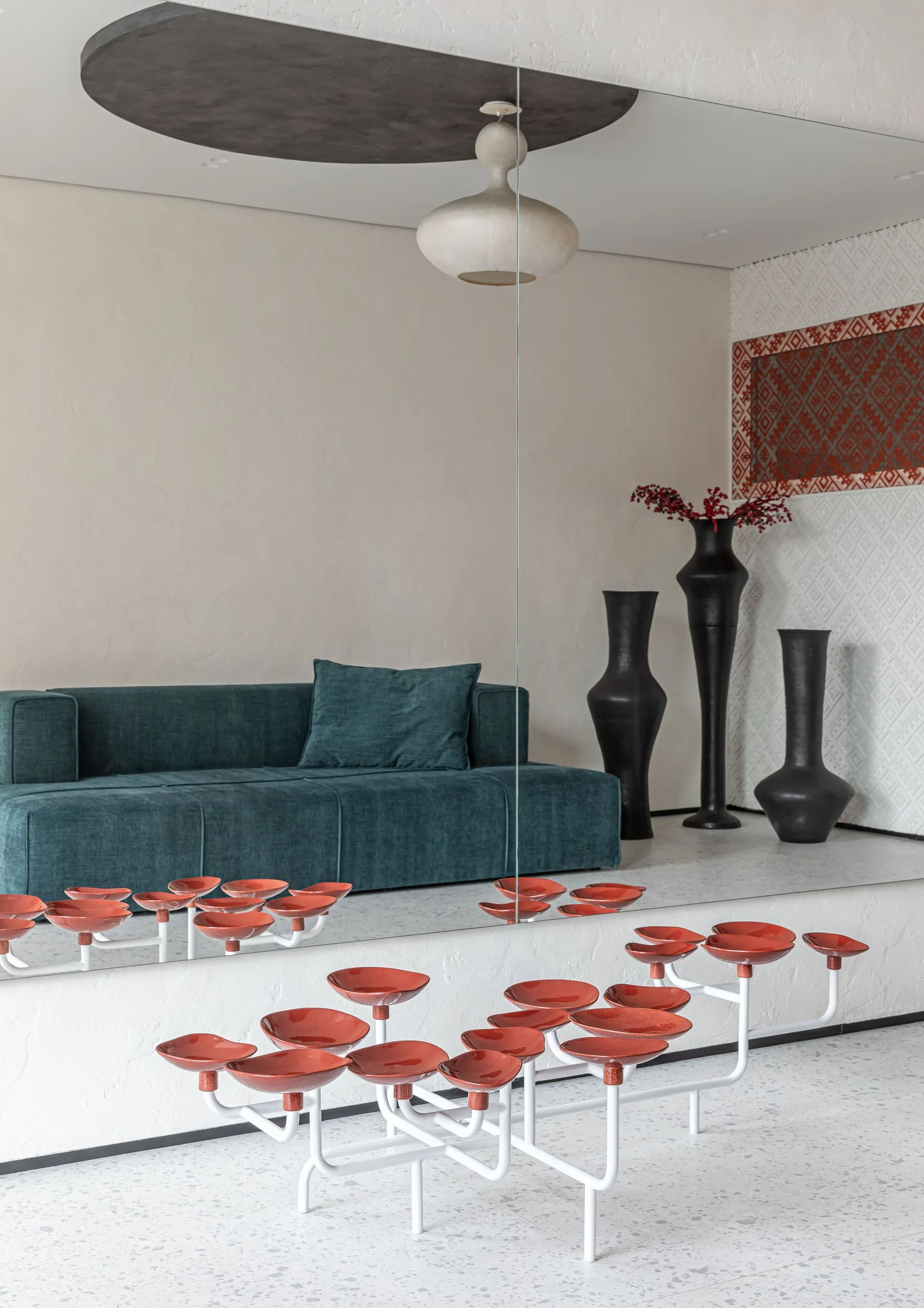 A modern living room featuring a deep teal sofa, a large wall mirror, three tall black floor vases, and a unique white floor fixture with red bowls.