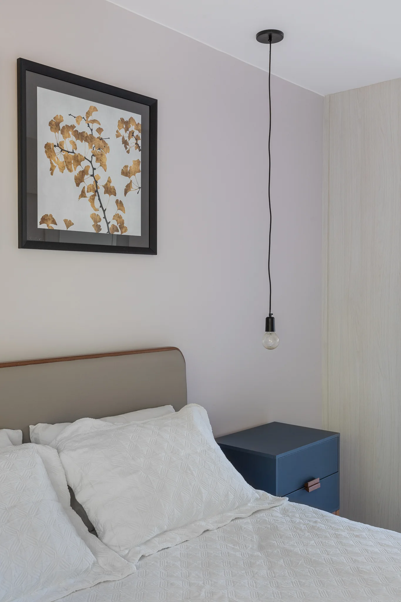A modern bedroom vignette featuring a bed with white textured bedding, a dark blue nightstand, a hanging bulb pendant, and framed ginkgo leaf artwork.