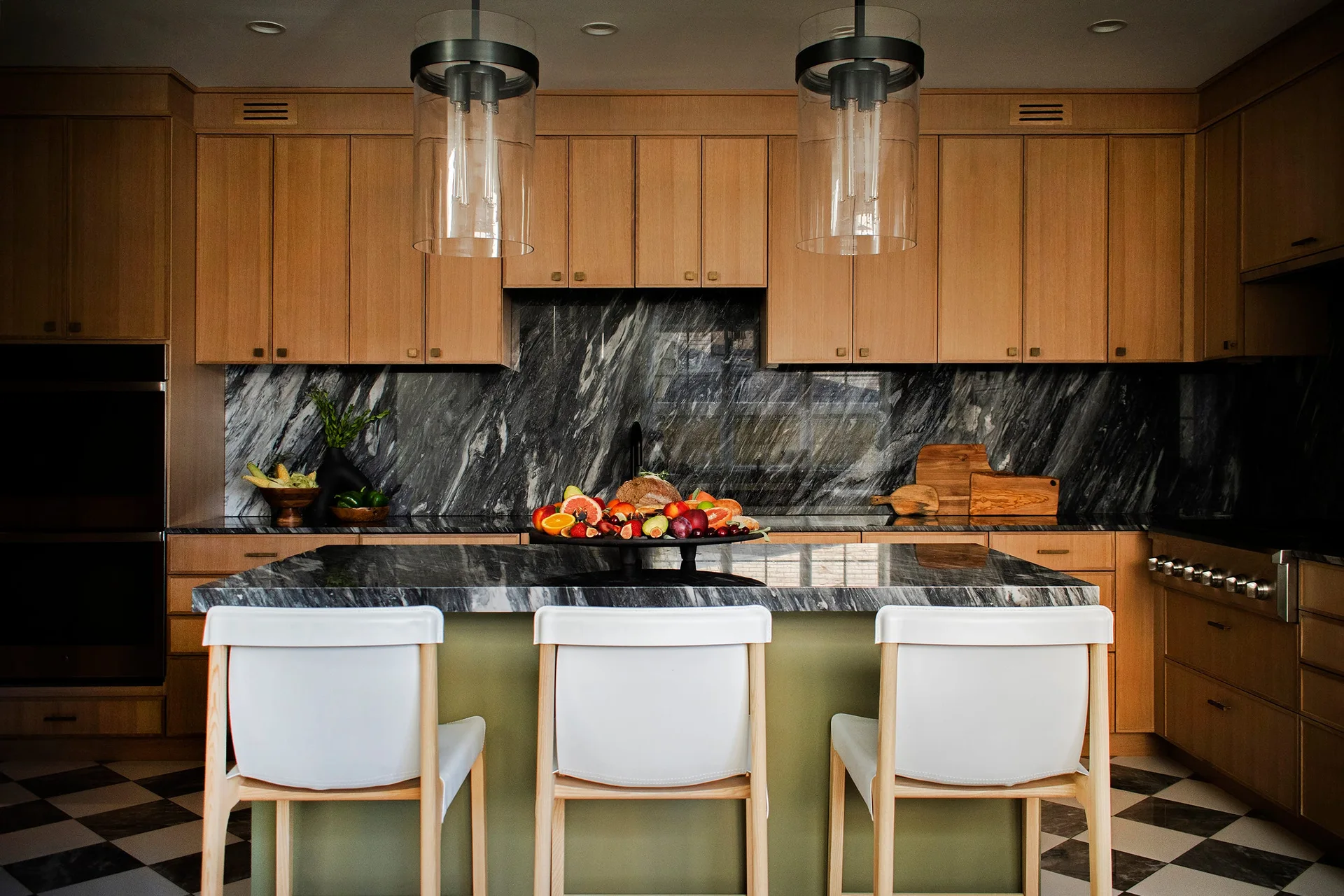 A modern kitchen featuring warm wood cabinets, a dark veined marble backsplash, a central olive green island with white bar stools, and checkered floors.
