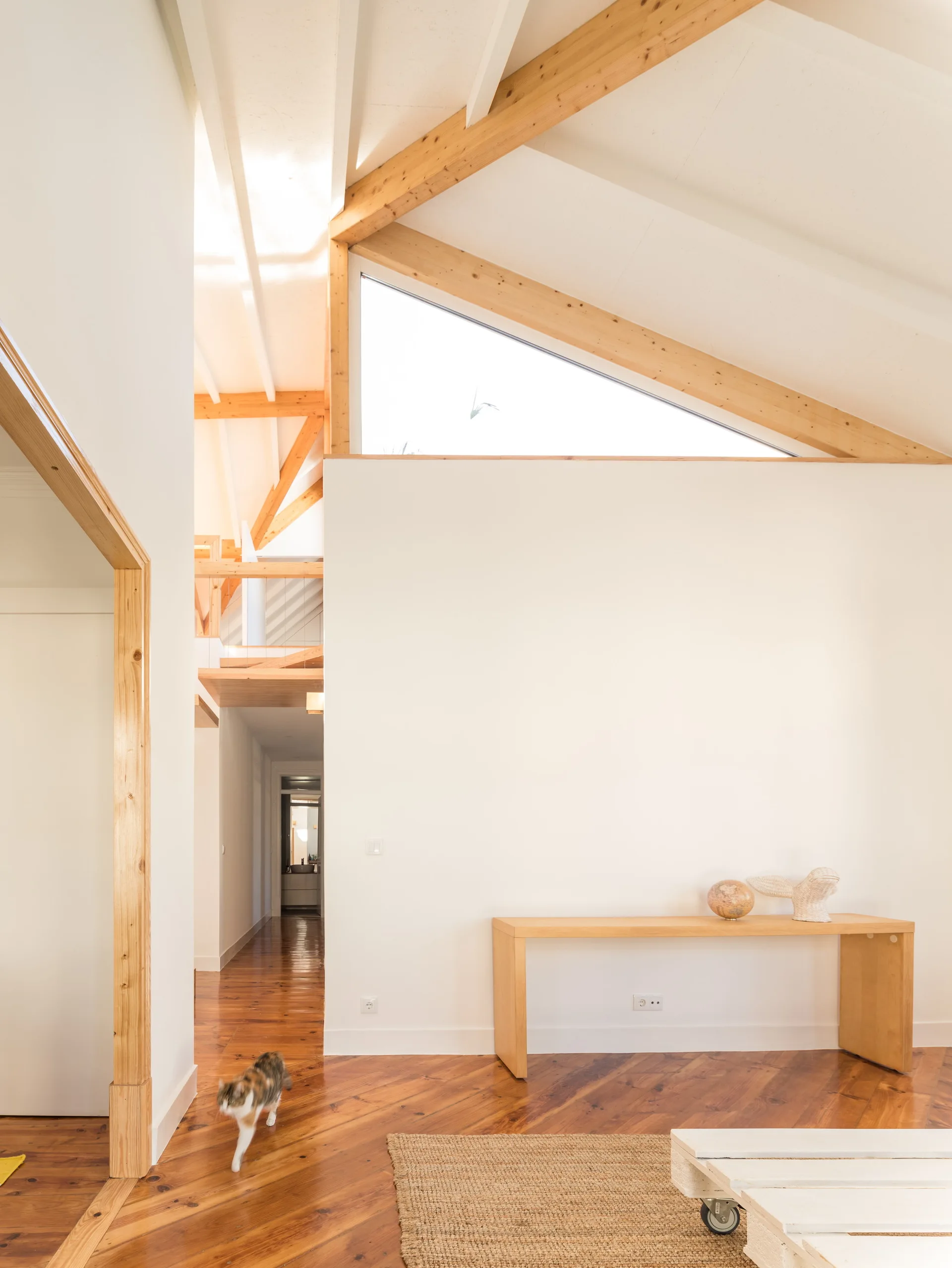 A bright living space features white walls, a vaulted ceiling with exposed timber beams, a triangular window, and a wooden console table.