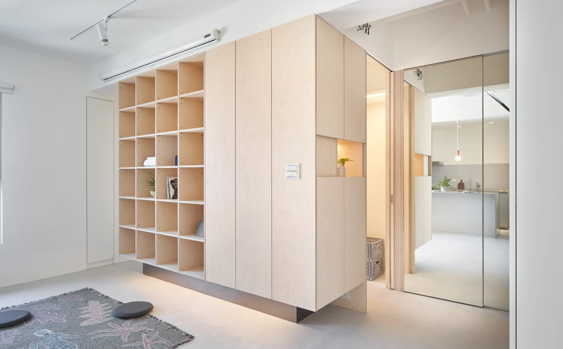 A light wood plywood storage unit with open shelves and closed cabinets stands in a minimalist living room next to a mirrored door.