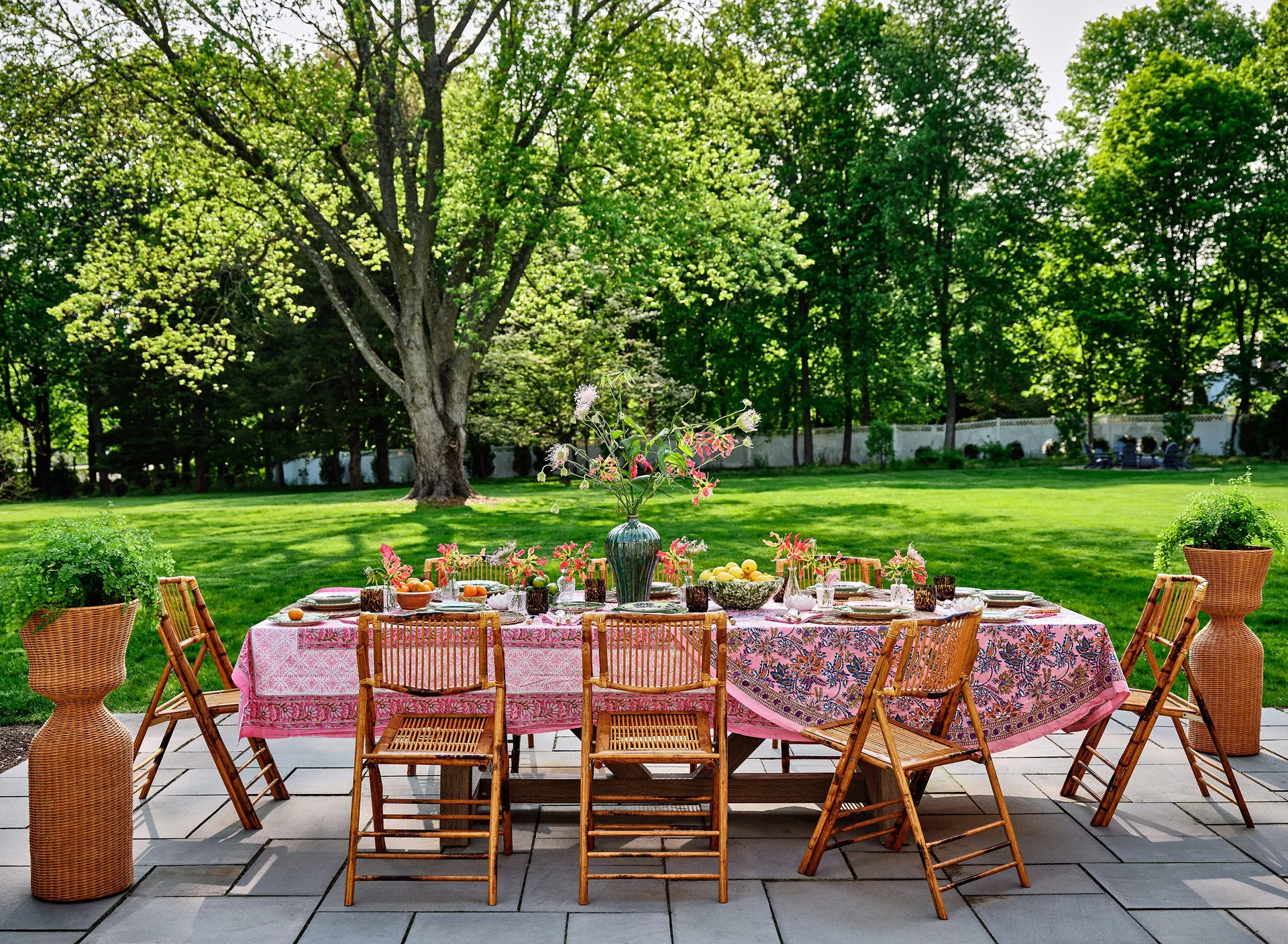 An outdoor dining table set with a pink floral tablecloth and bamboo folding chairs on a stone patio overlooking a lush green lawn.