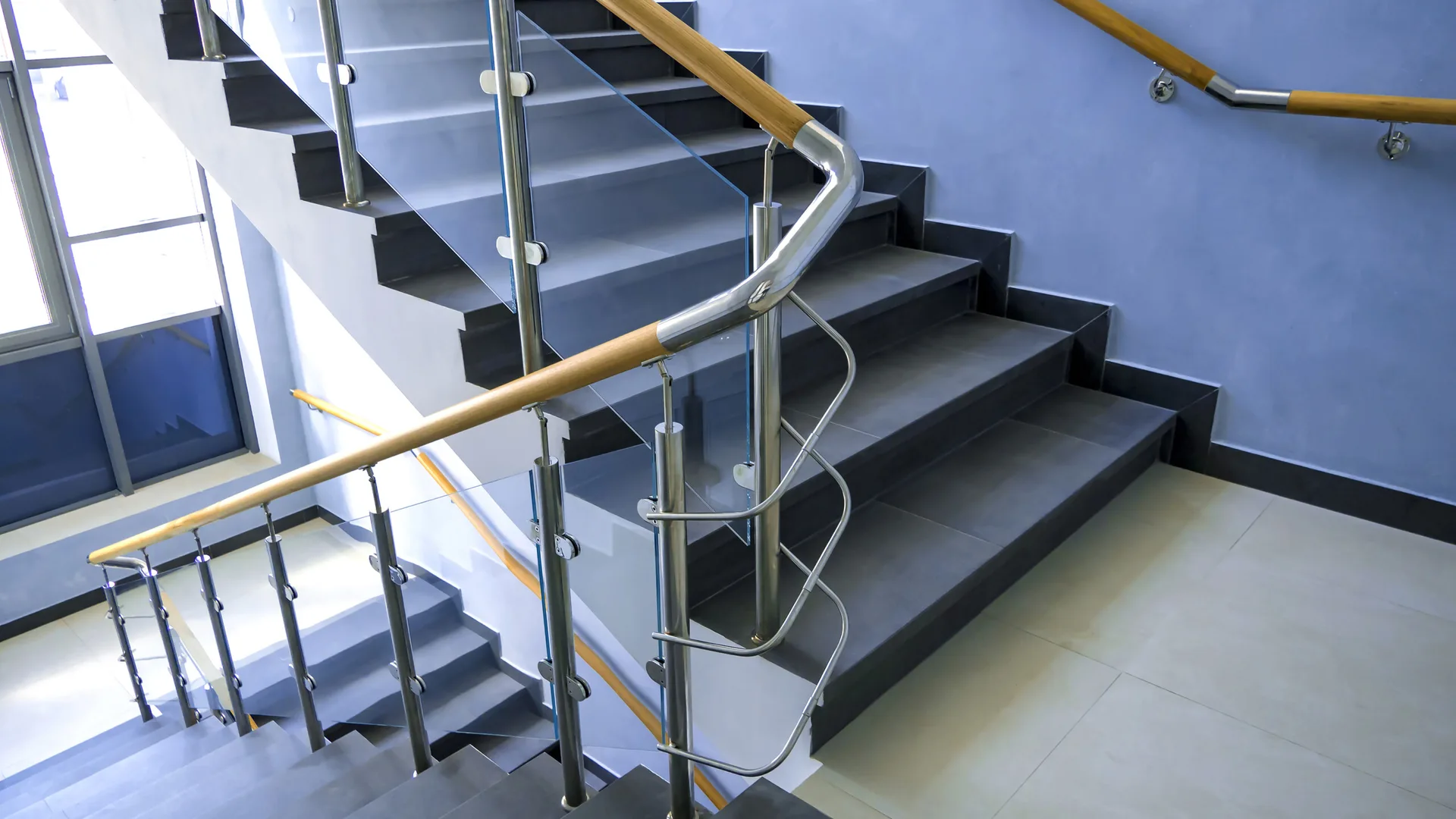 A modern multi-level staircase with dark grey tiled steps, glass and stainless steel railings, and wooden handrail details.