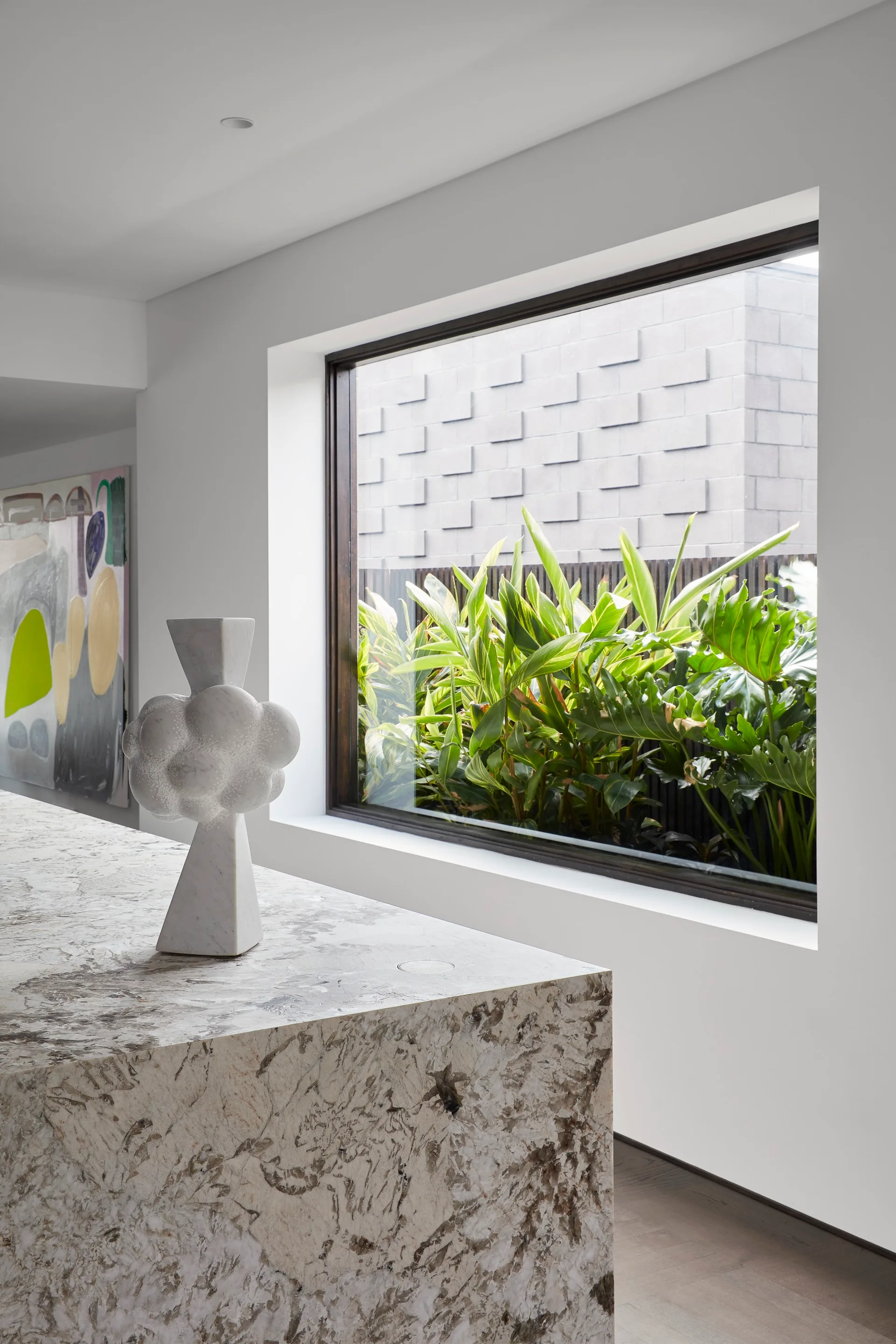 A modern kitchen island with a veined stone countertop features a white abstract sculpture next to a picture window showcasing green garden plants.