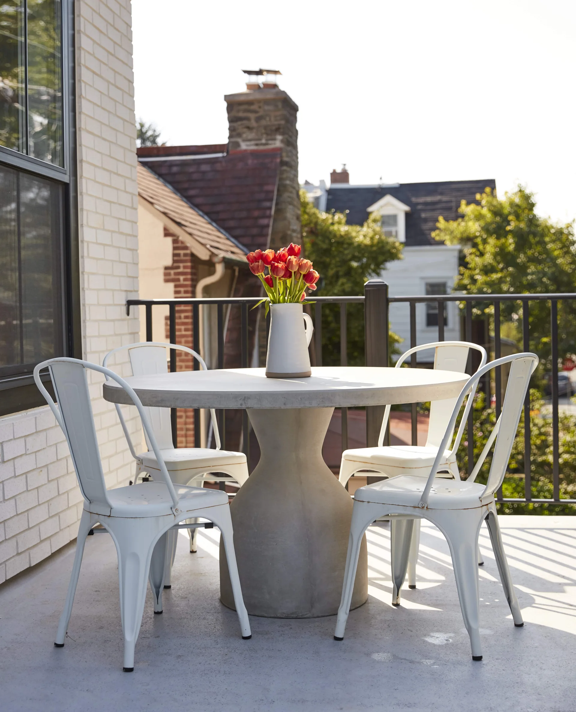 Four white metal bistro chairs surround a round concrete pedestal table topped with red tulips on a sunny balcony with a painted white brick wall.