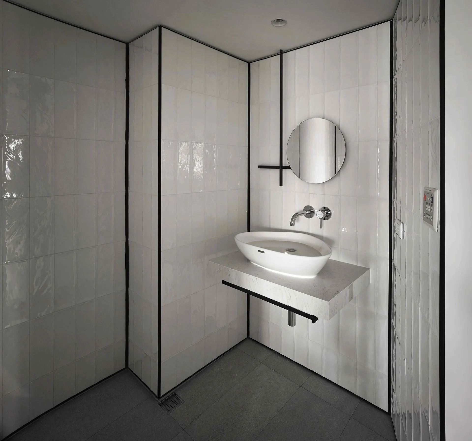 A minimalist bathroom corner features high-gloss white wall tiles with sharp black trim, a floating stone vanity, and an oval vessel sink.