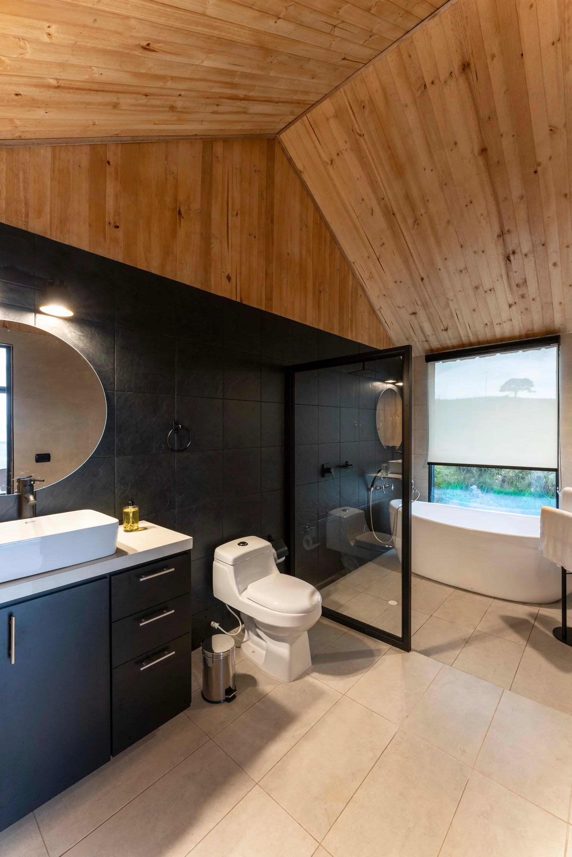 A modern bathroom with a wood-paneled vaulted ceiling features a black tiled wall, a dark vanity, a white toilet, and a freestanding bathtub near a window.