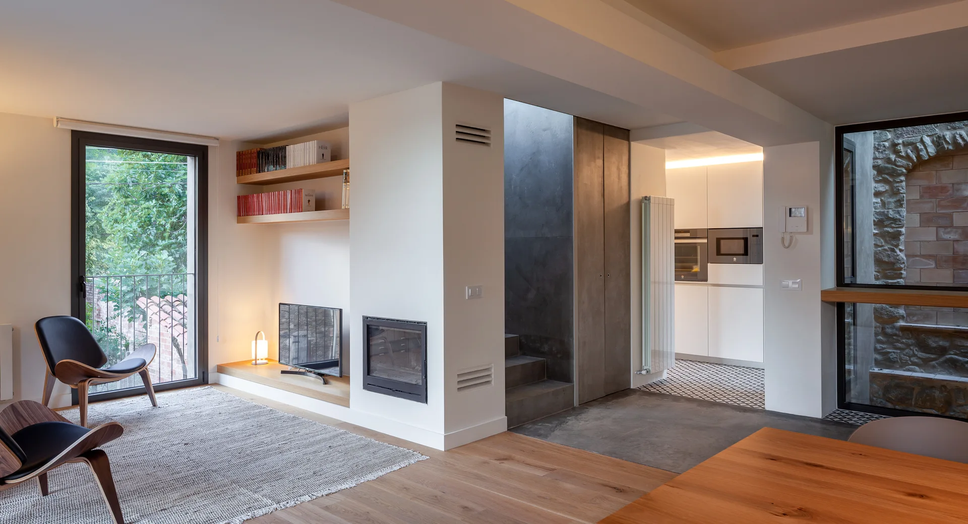 A minimalist living space featuring wood lounge chairs, a built-in fireplace column, floating shelves, and concrete stairs leading to a kitchen.