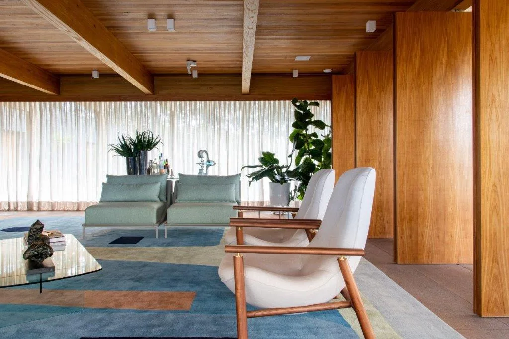 A modern living room with warm wood paneling on the walls and ceiling, white wooden armchairs, and a large blue geometric area rug.