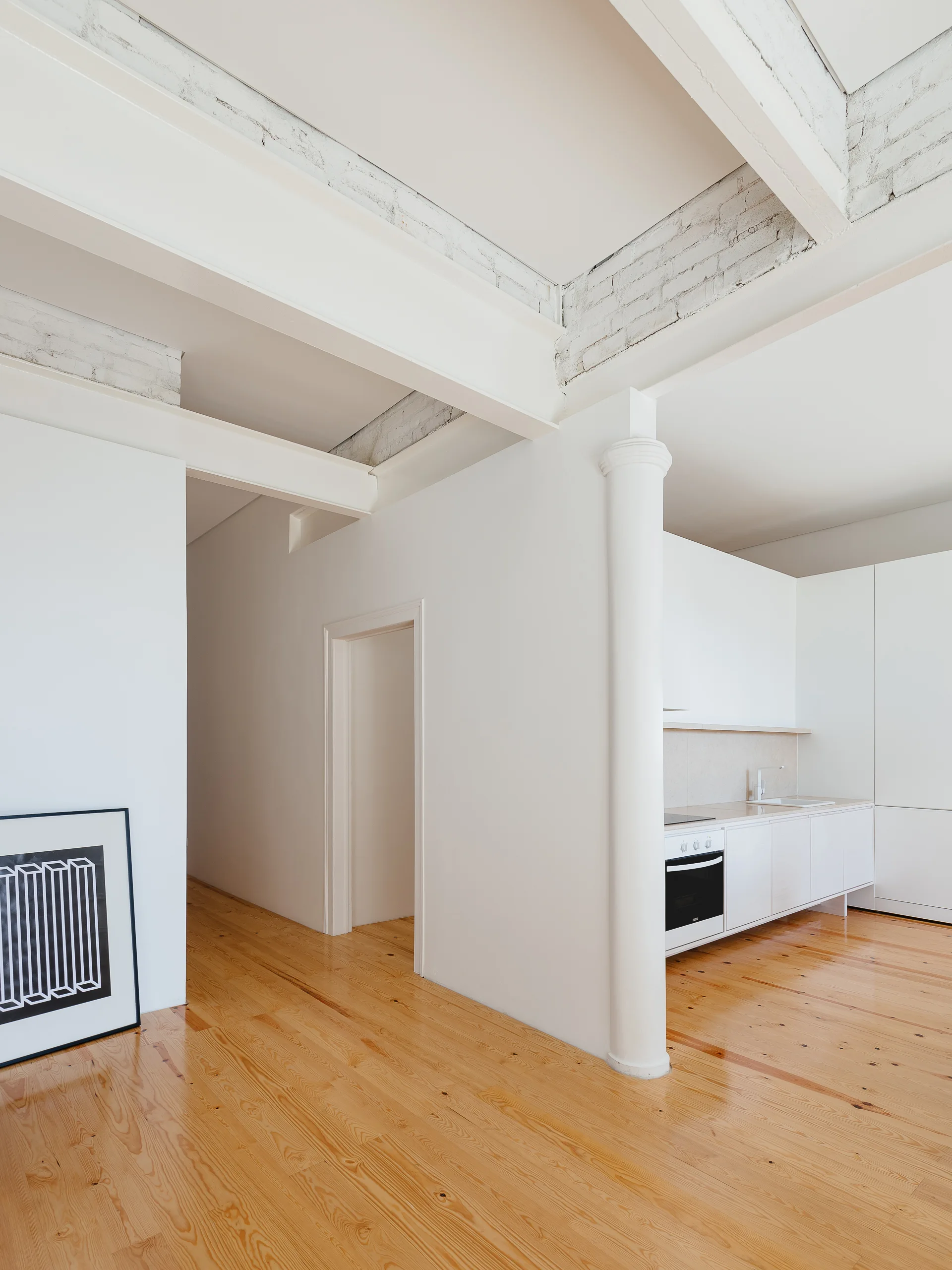 A minimalist white kitchen features flat cabinets set against pine wood floors and a painted beam ceiling with a structural column.