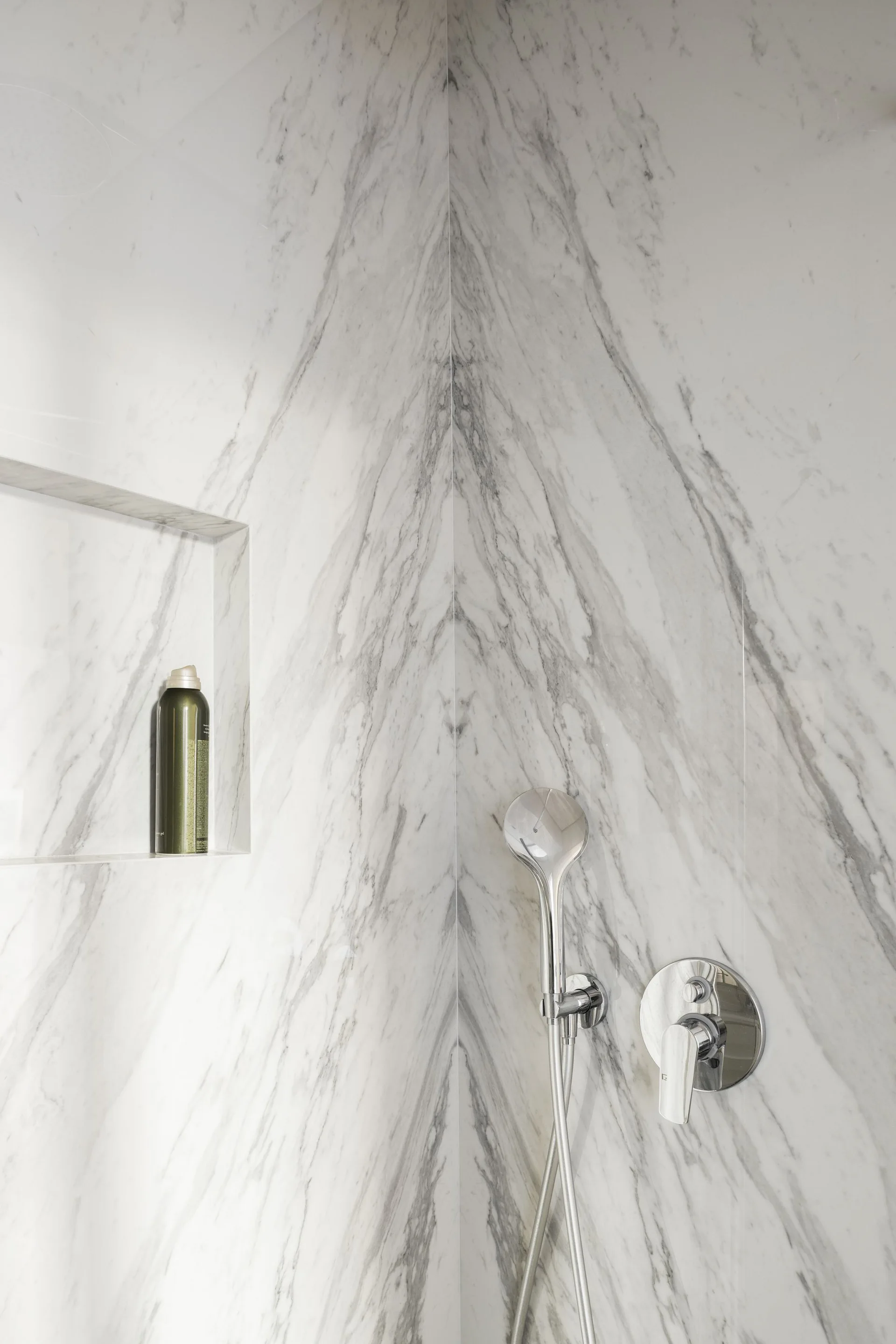 A close-up of a bathroom shower corner featuring bookmatched white marble tiles, a recessed shelf, and chrome fixtures.