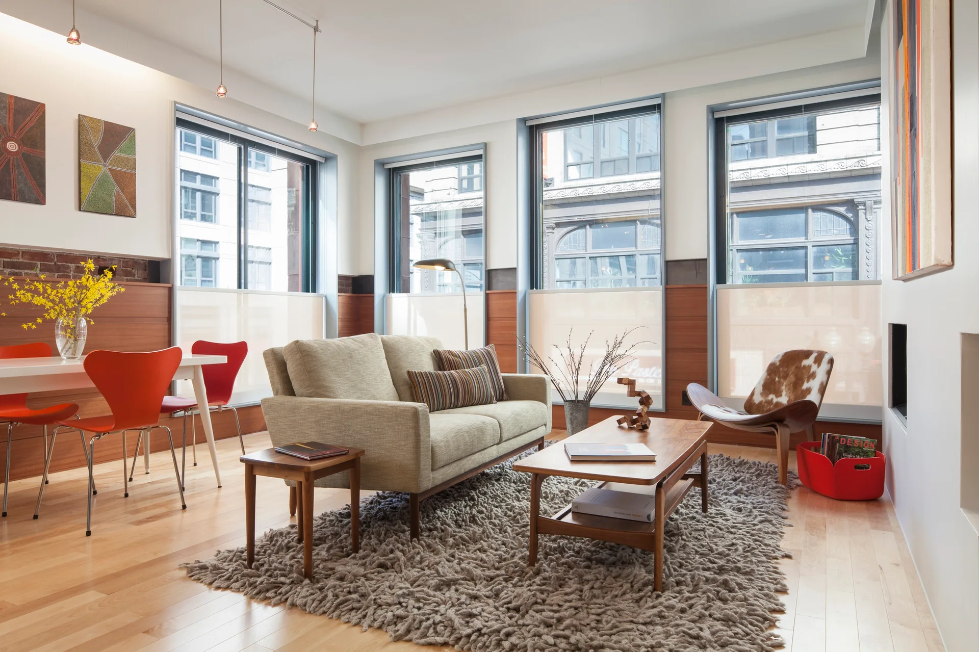A mid-century modern living room features a cream sofa, textured grey shag rug, and orange dining chairs in an open layout with horizontal wood wainscoting.