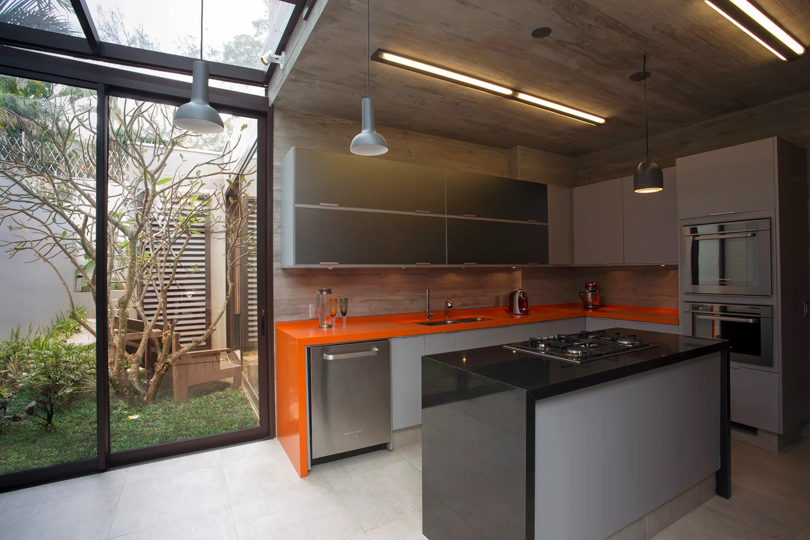 A modern kitchen with bright orange countertops, gray cabinetry, a dark island, and a sliding glass door opening to a garden.
