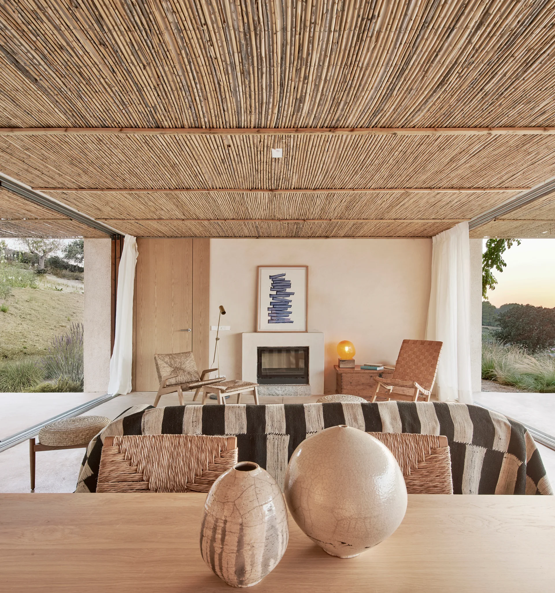 An organic modern living room featuring a textured reed ceiling, a central fireplace, a striped sofa, and large sliding glass doors.