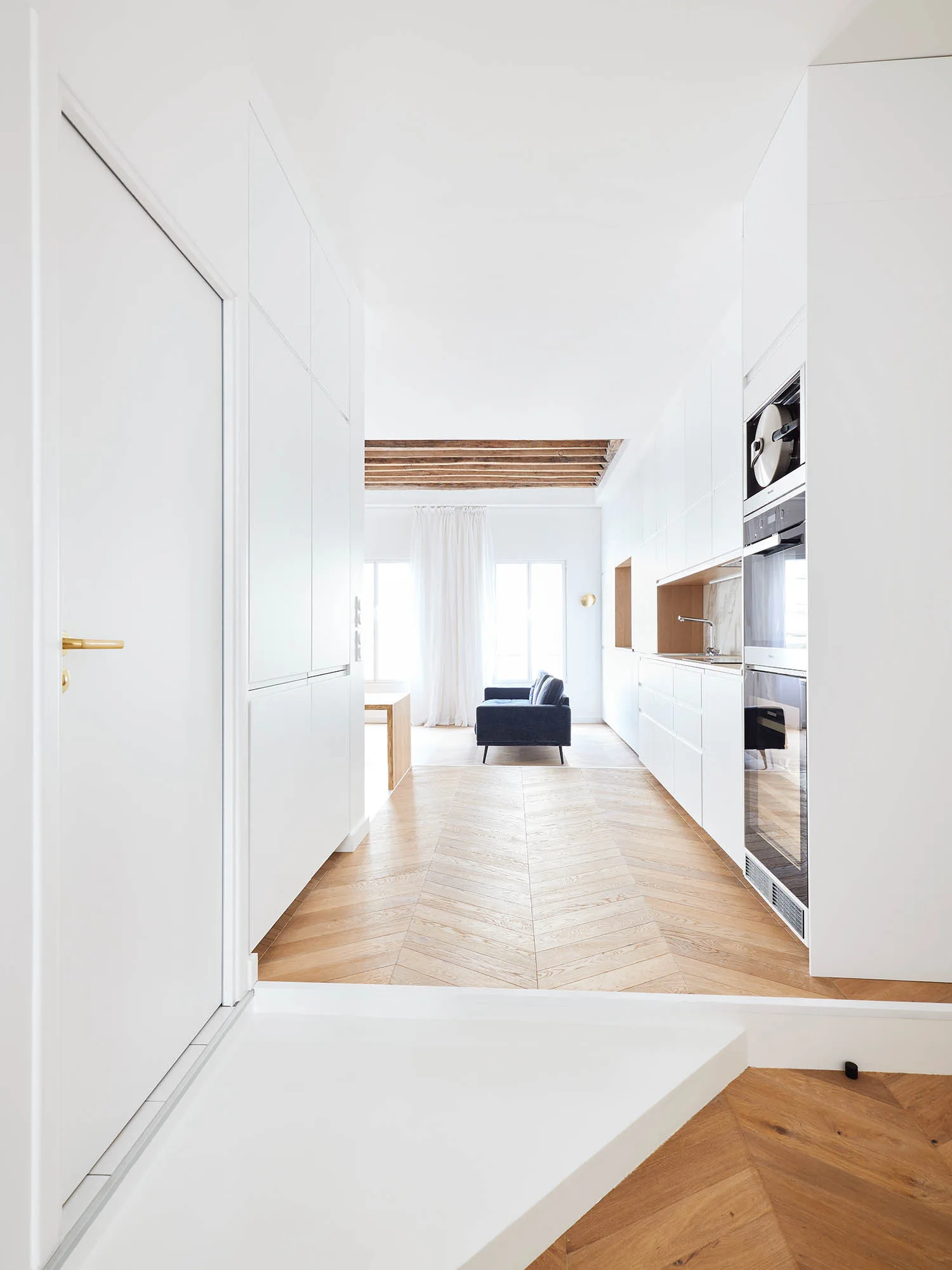 A white galley kitchen features integrated flat-front cabinets, built-in appliances, and light wood herringbone parquet flooring leading to a living area.