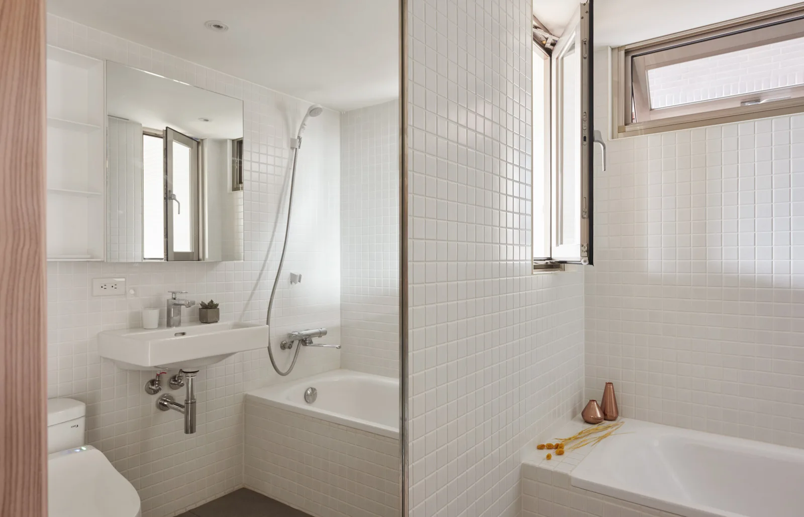 A bright minimalist bathroom features white square grid tile walls, a wall-mounted ceramic sink, a mirror, and a recessed bathtub.