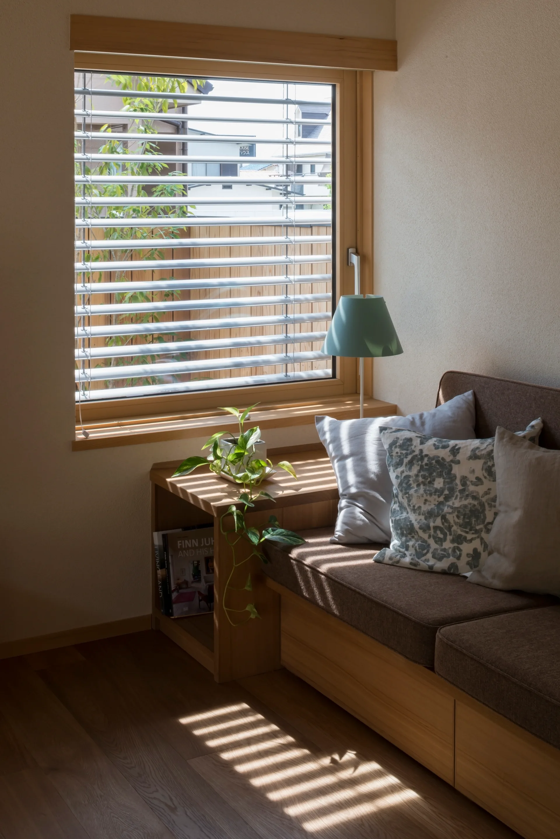 A built-in wooden bench with brown upholstery sits below a window with horizontal blinds casting striped shadows across the wood floor.
