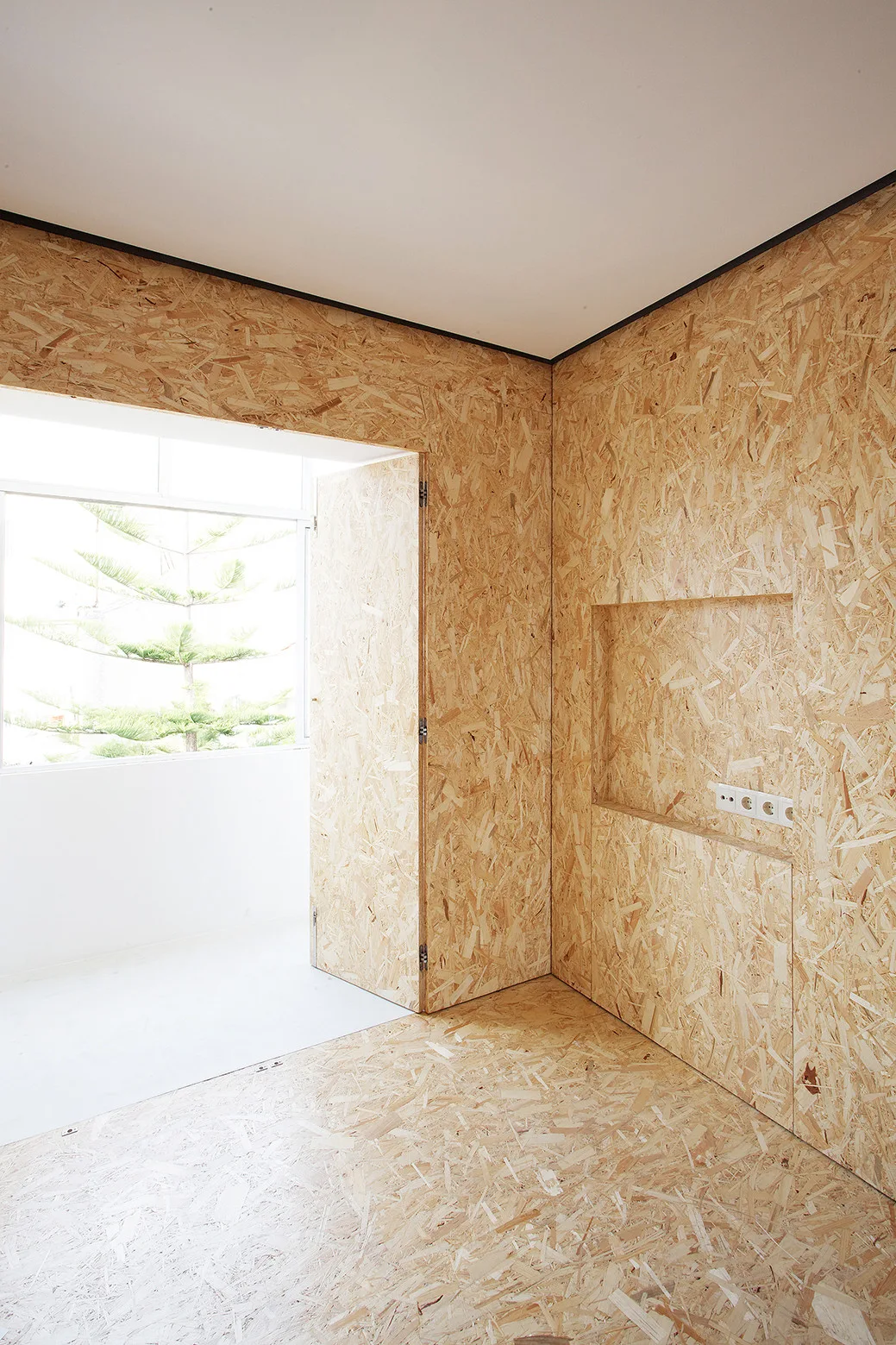 An interior space features walls and floors clad in textured oriented strand board panels, with a recessed niche and an open doorway.