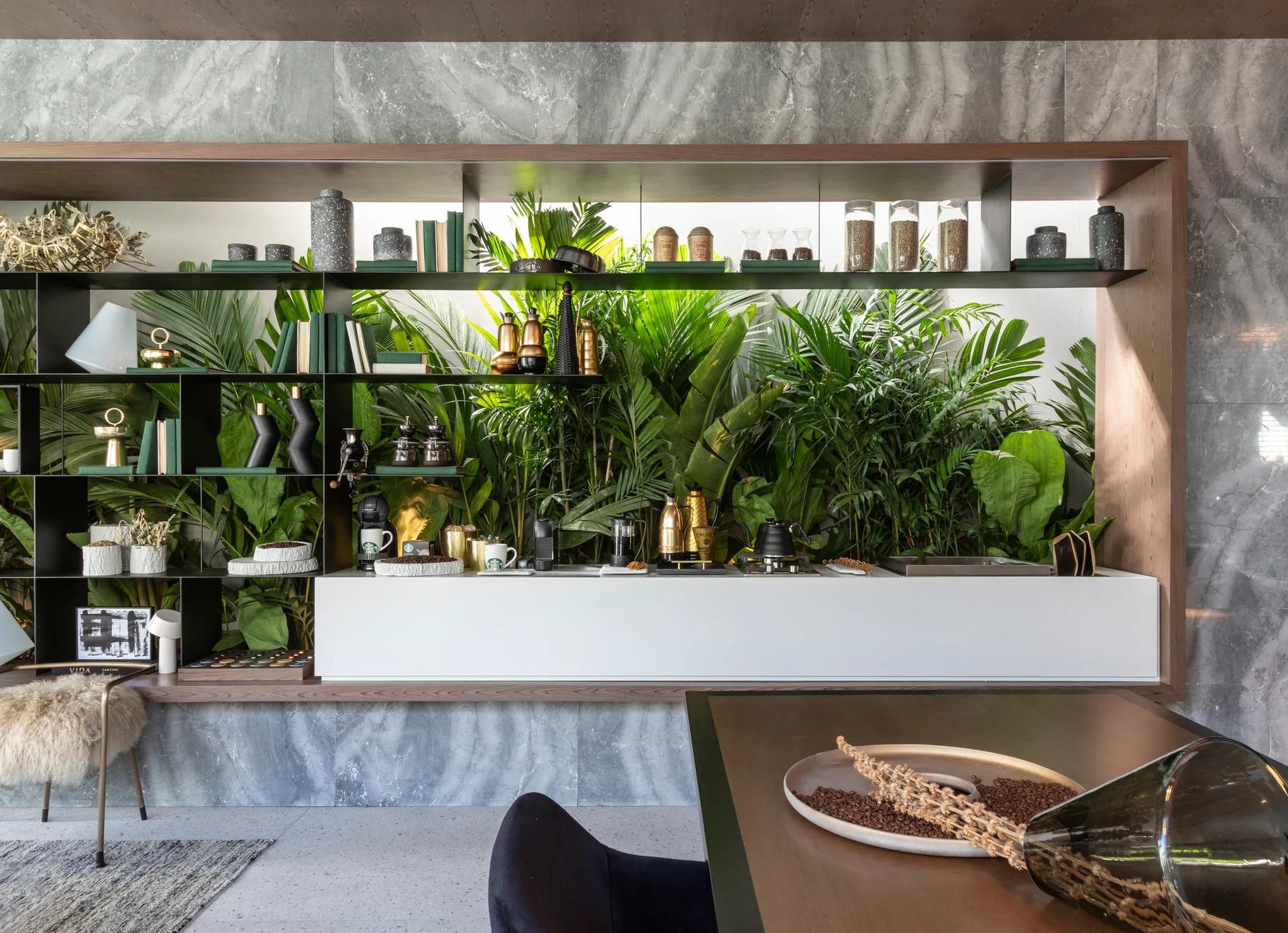 A built-in white coffee station with black shelving and lush green plants is set against a grey marbled stone wall in a modern dining area.