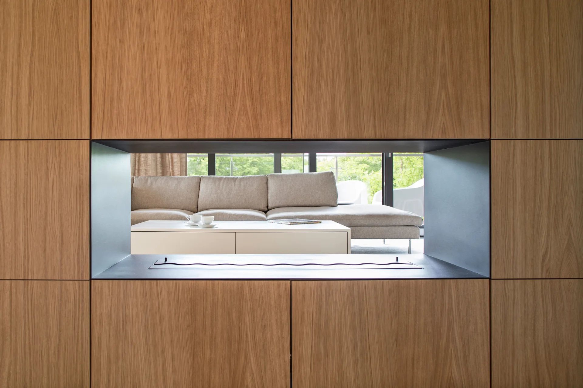 A rectangular see-through fireplace opening in a wood-paneled wall reveals a modern living room with a grey sectional sofa and a white coffee table.