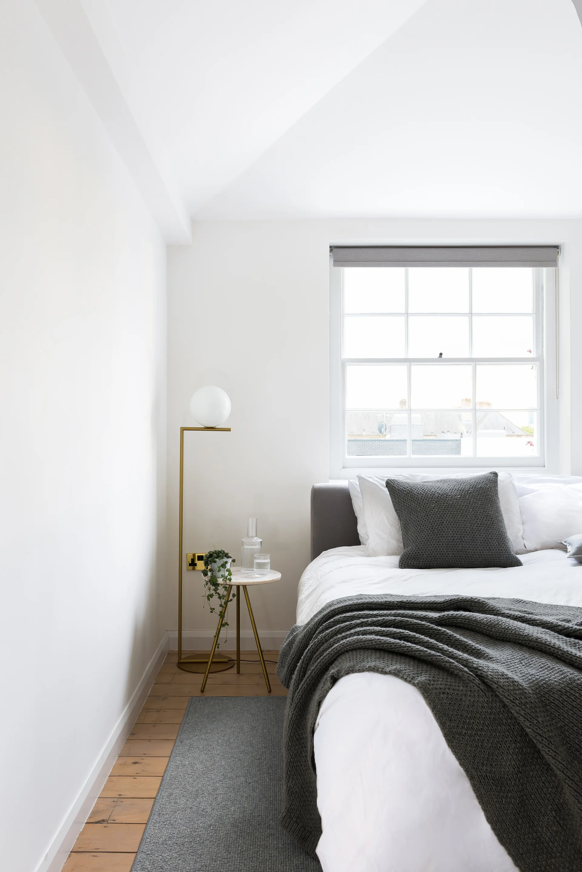 A minimalist bedroom features a bed with grey knit bedding, a slim brass floor lamp, and a small marble-topped side table on wood flooring.