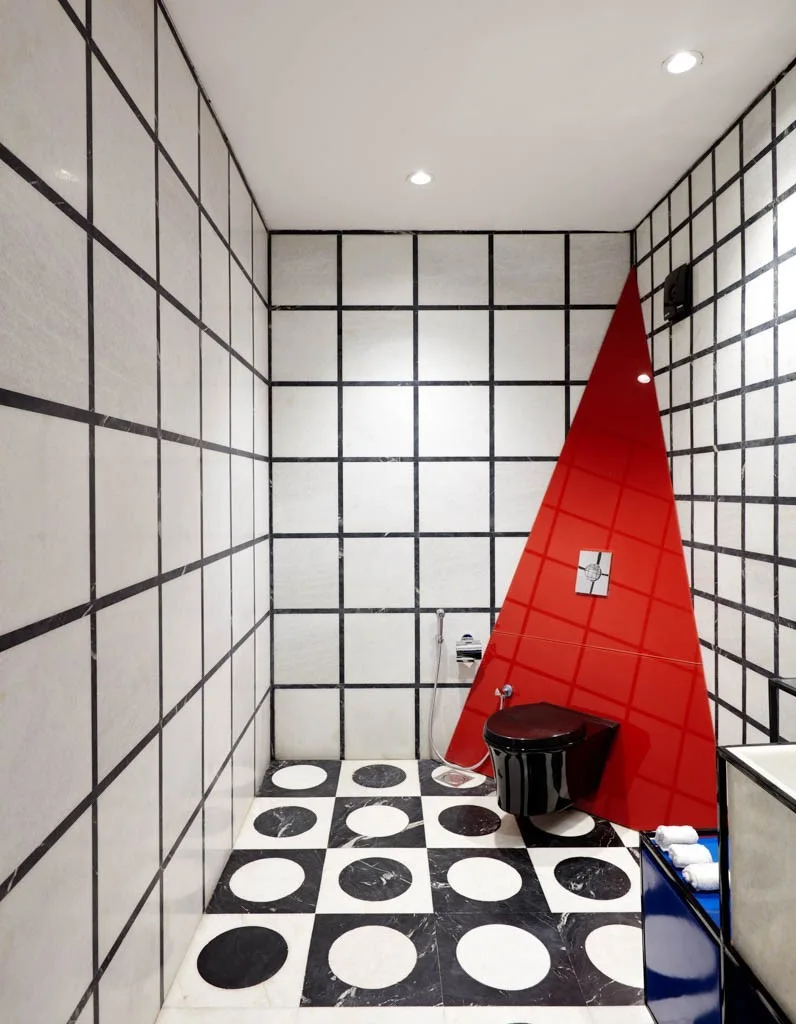 A graphic bathroom features white grid wall tiles, a black and white geometric patterned floor, and a bold red triangular panel behind a black toilet.