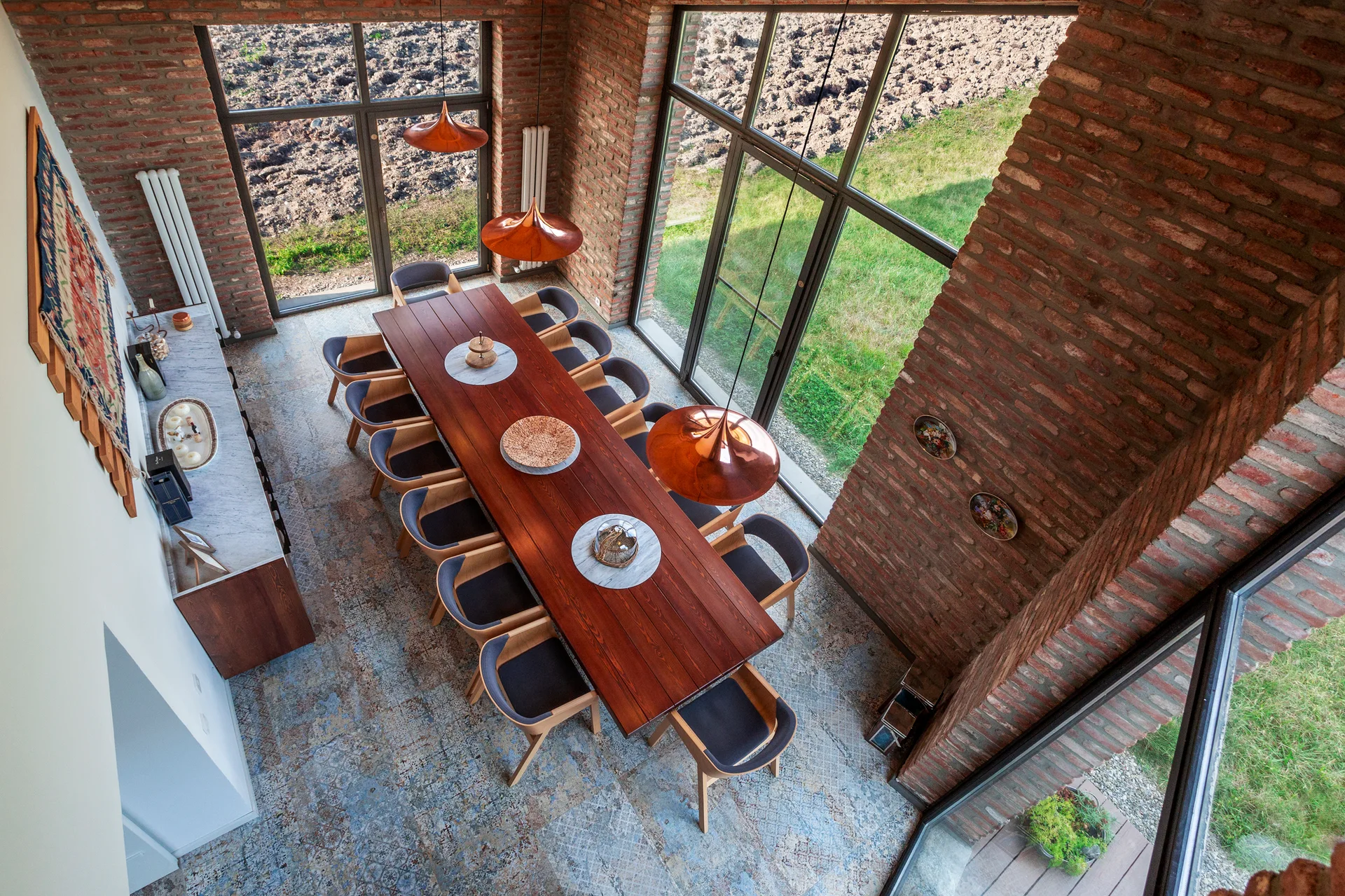 An overhead view of a long wooden dining table and chairs on patterned tile floor between exposed brick walls and large floor-to-ceiling windows.