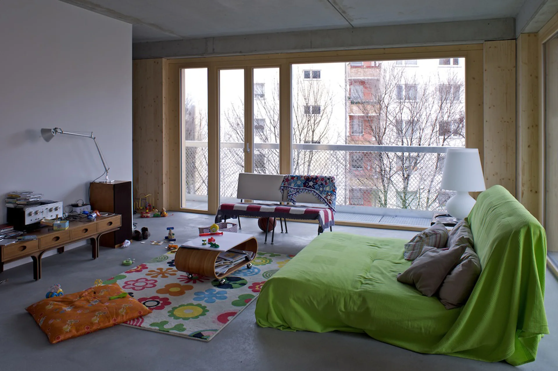 A casual living room features an eclectic mix of furniture, a green covered sofa, a floral pattern rug, and floor-to-ceiling windows.