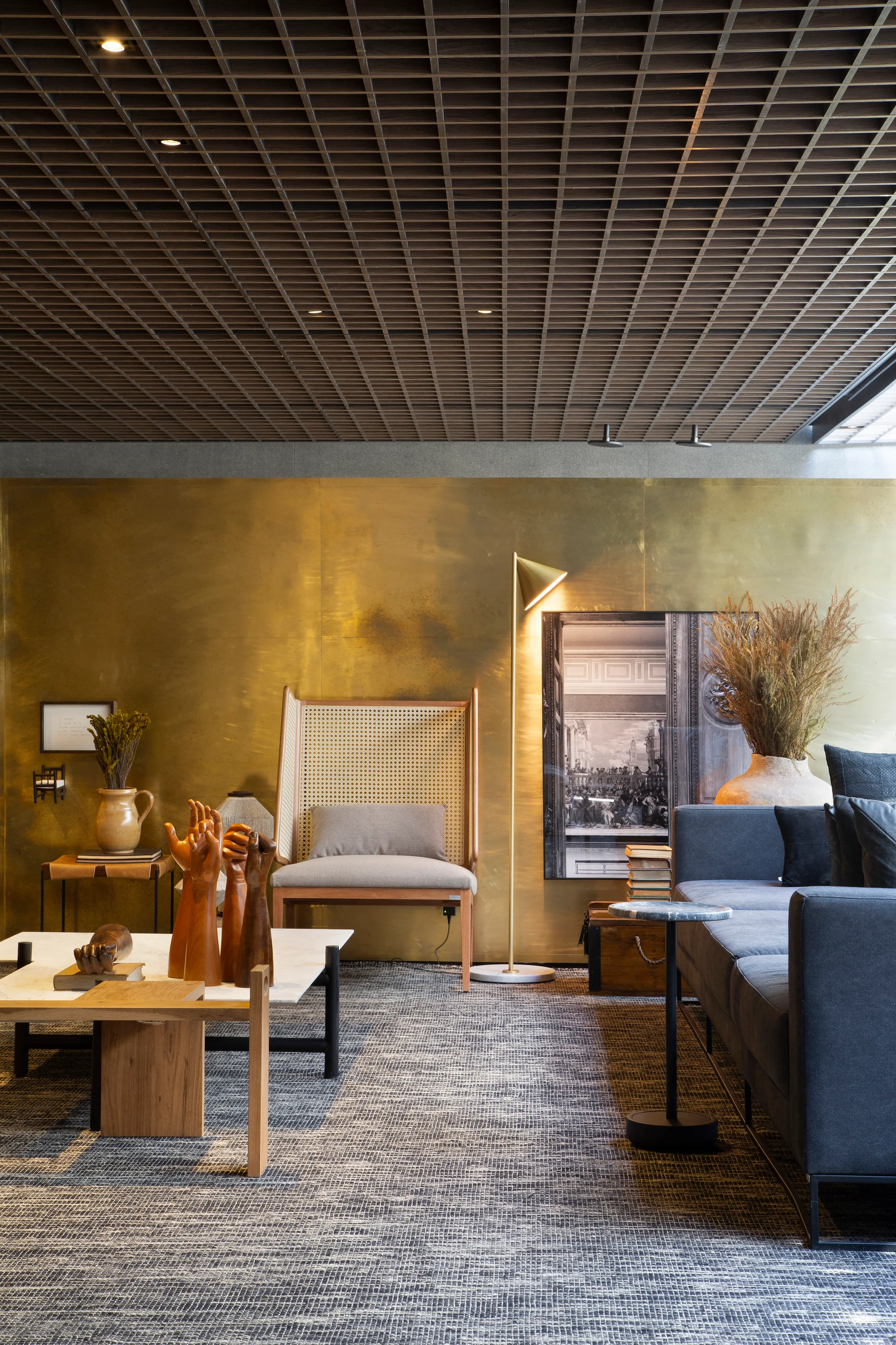 A contemporary living room features a brushed brass wall panel, a dark wood grid ceiling, a grey sofa, and a cane-back armchair.