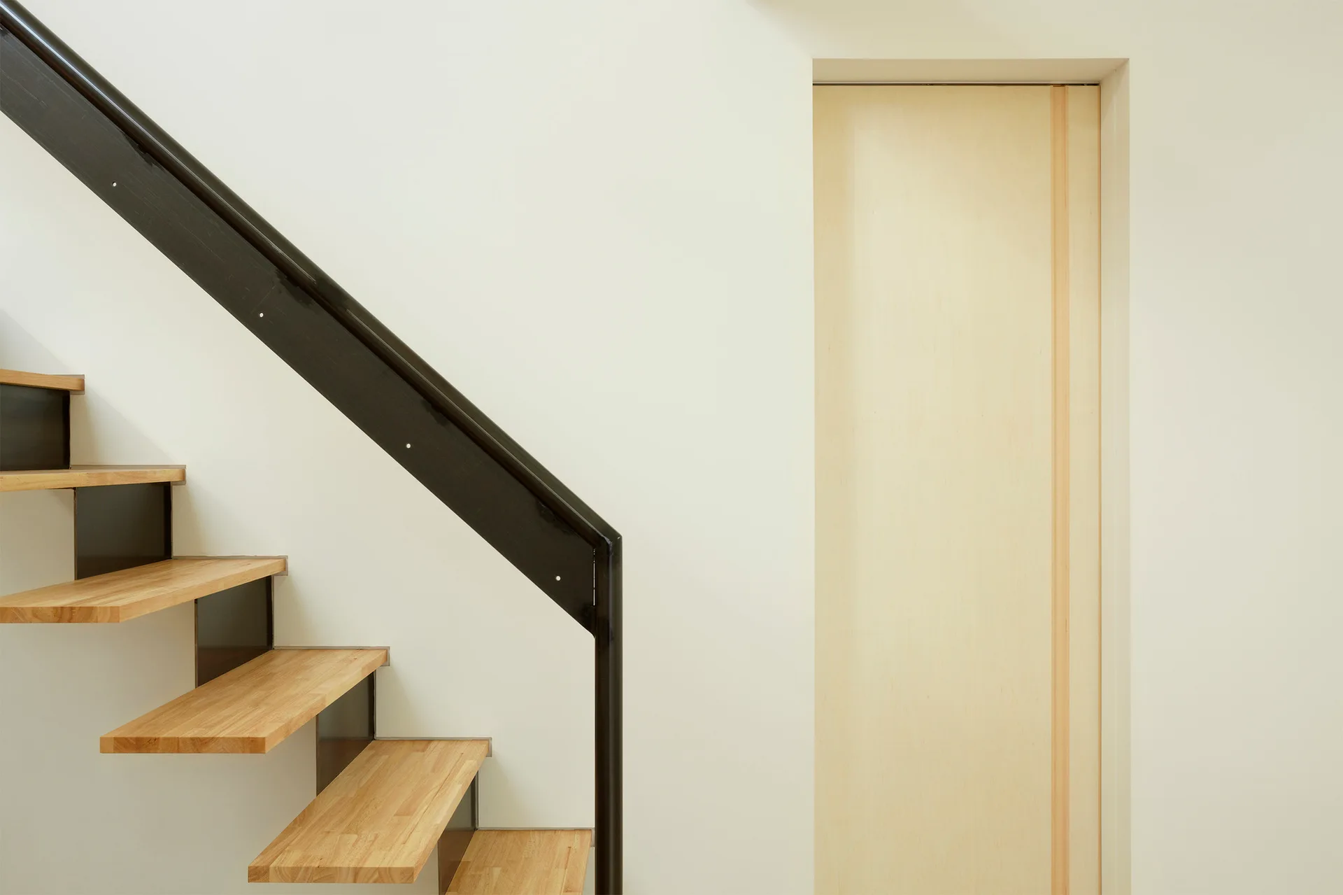 A minimalist open-riser wooden staircase with a black metal handrail stands next to a flush light wood door set into a white wall.