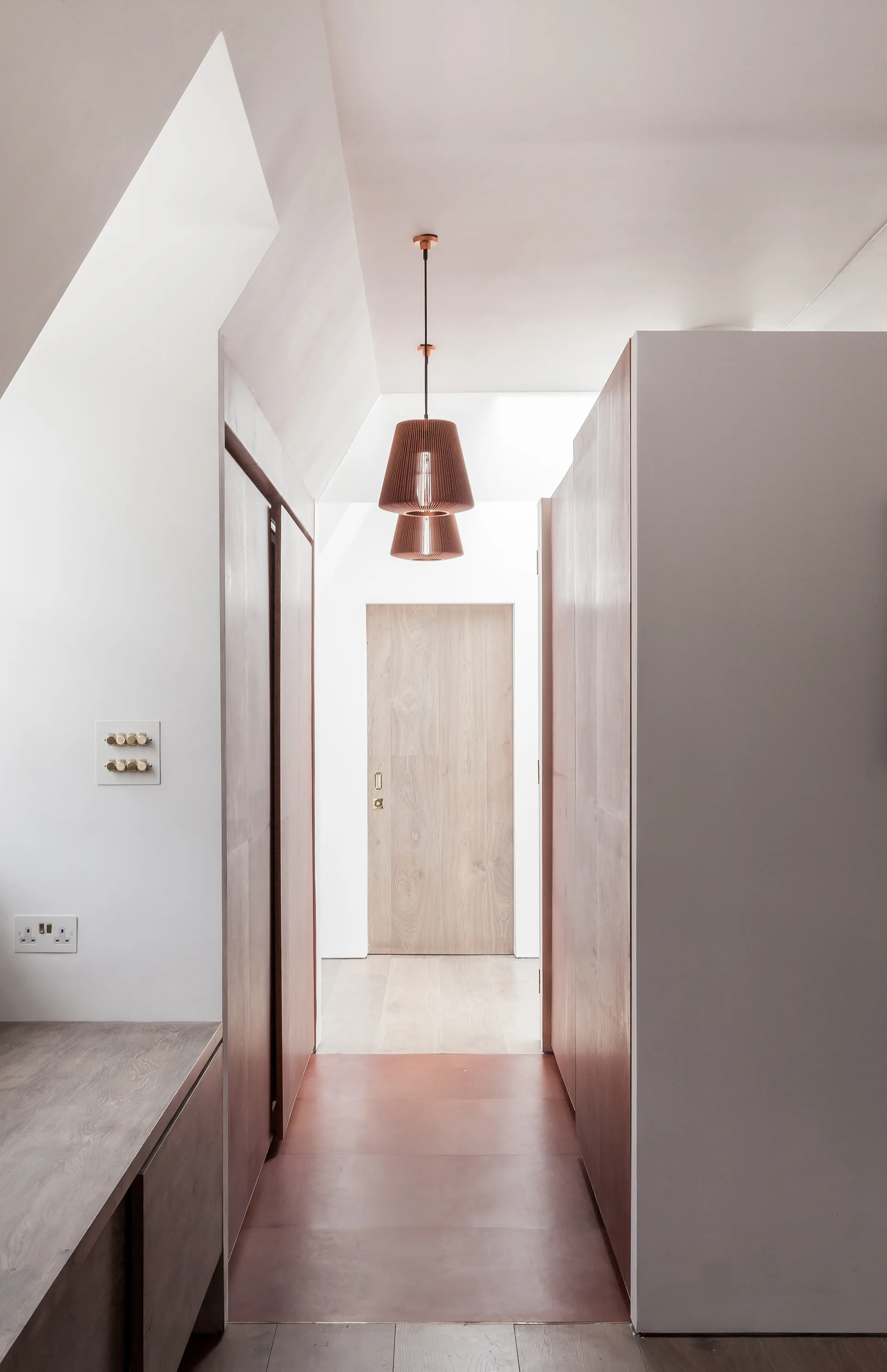 A minimalist hallway with wood-paneled walls, a flush light-wood door at the far end, and tiered copper pendant lights hanging from the ceiling.
