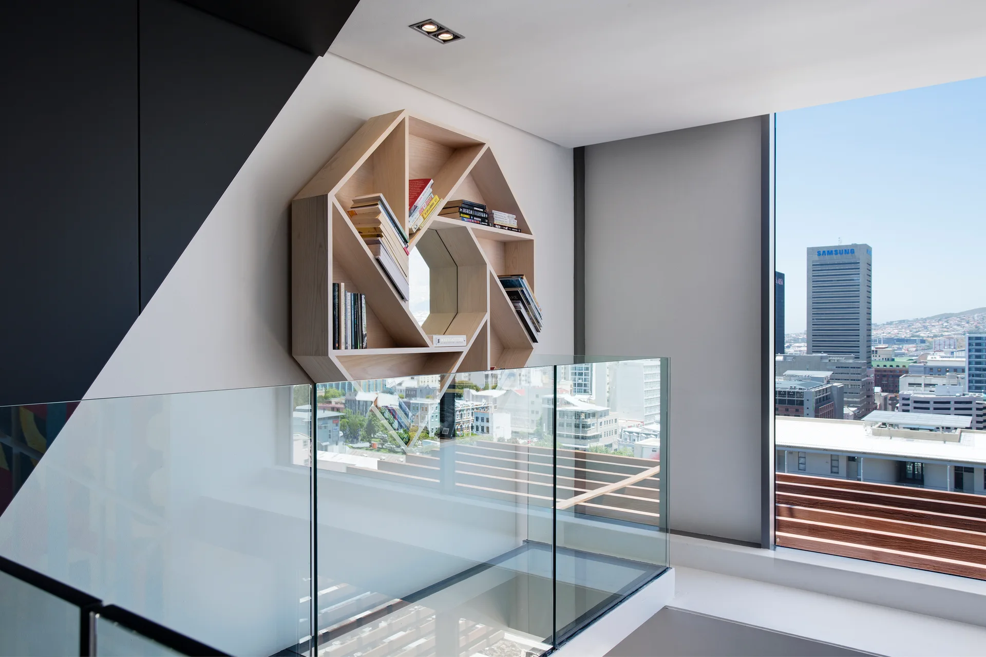 An indoor stair landing features an octagonal light wood wall shelf filled with books, a clear glass railing, and a window displaying a city view.