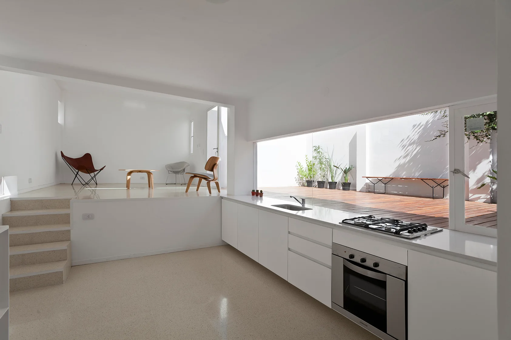 A minimalist kitchen features white lower cabinets below a long horizontal window looking onto a wood deck, with stairs leading to a raised living space.