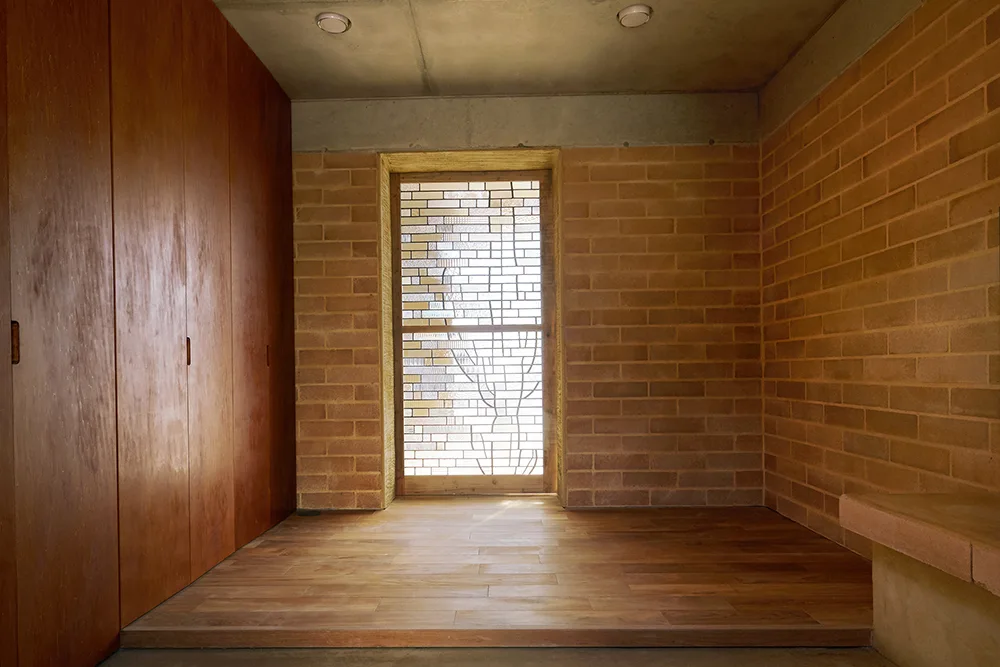 An entryway features terracotta brick walls, a concrete ceiling, a tall wood storage wall, and a raised wooden floor leading to a patterned glass door.