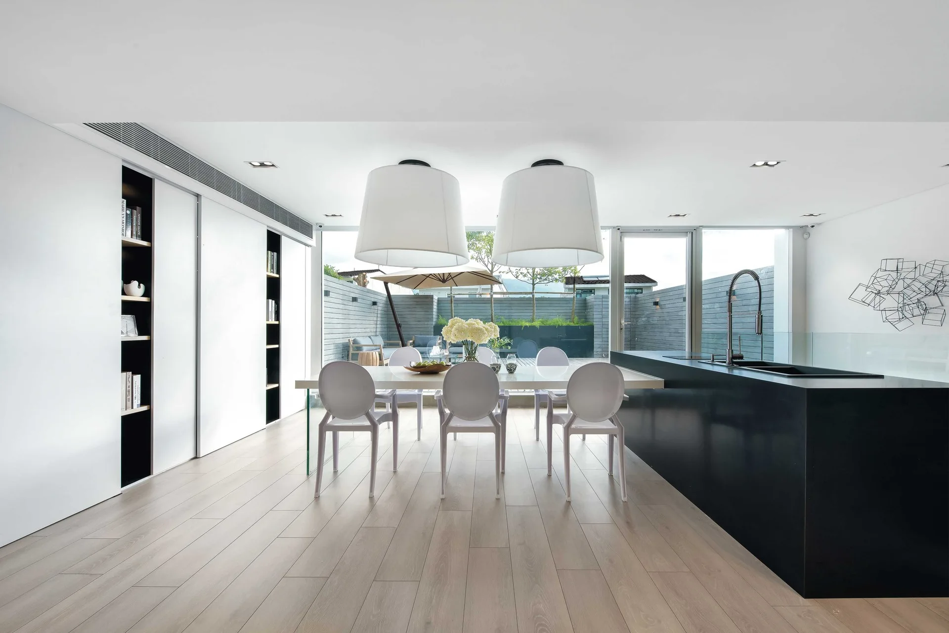 A minimalist dining area has a white table with molded chairs, large white pendant lights, and a black kitchen island near a sliding glass patio door.