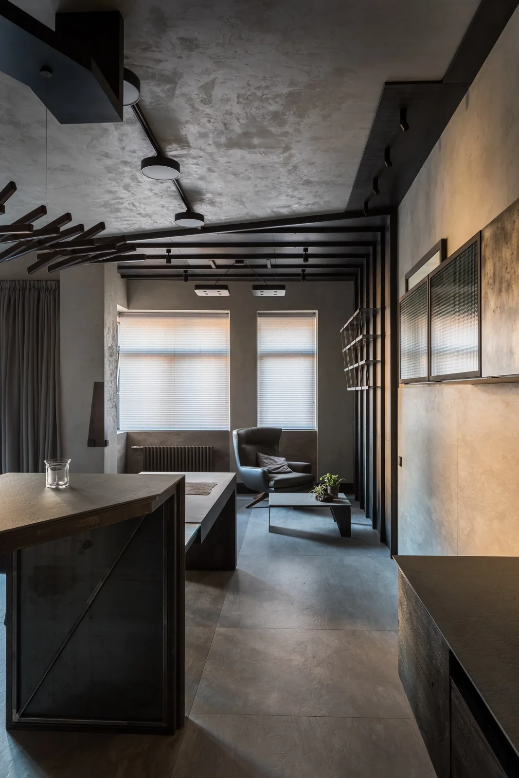 A moody industrial living room features textured grey walls and floors, black ceiling beams with track lights, and a dark leather armchair by the window.
