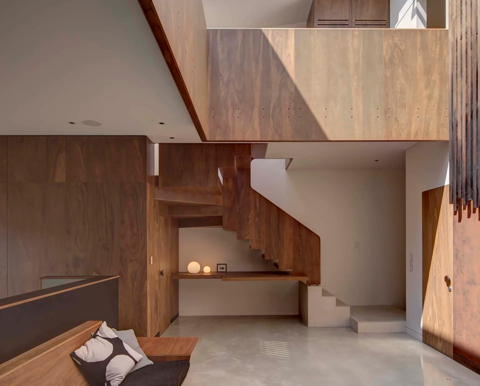A modern wooden staircase with rich walnut paneling sits above a smooth concrete floor next to a built-in floating desk and a low bench.