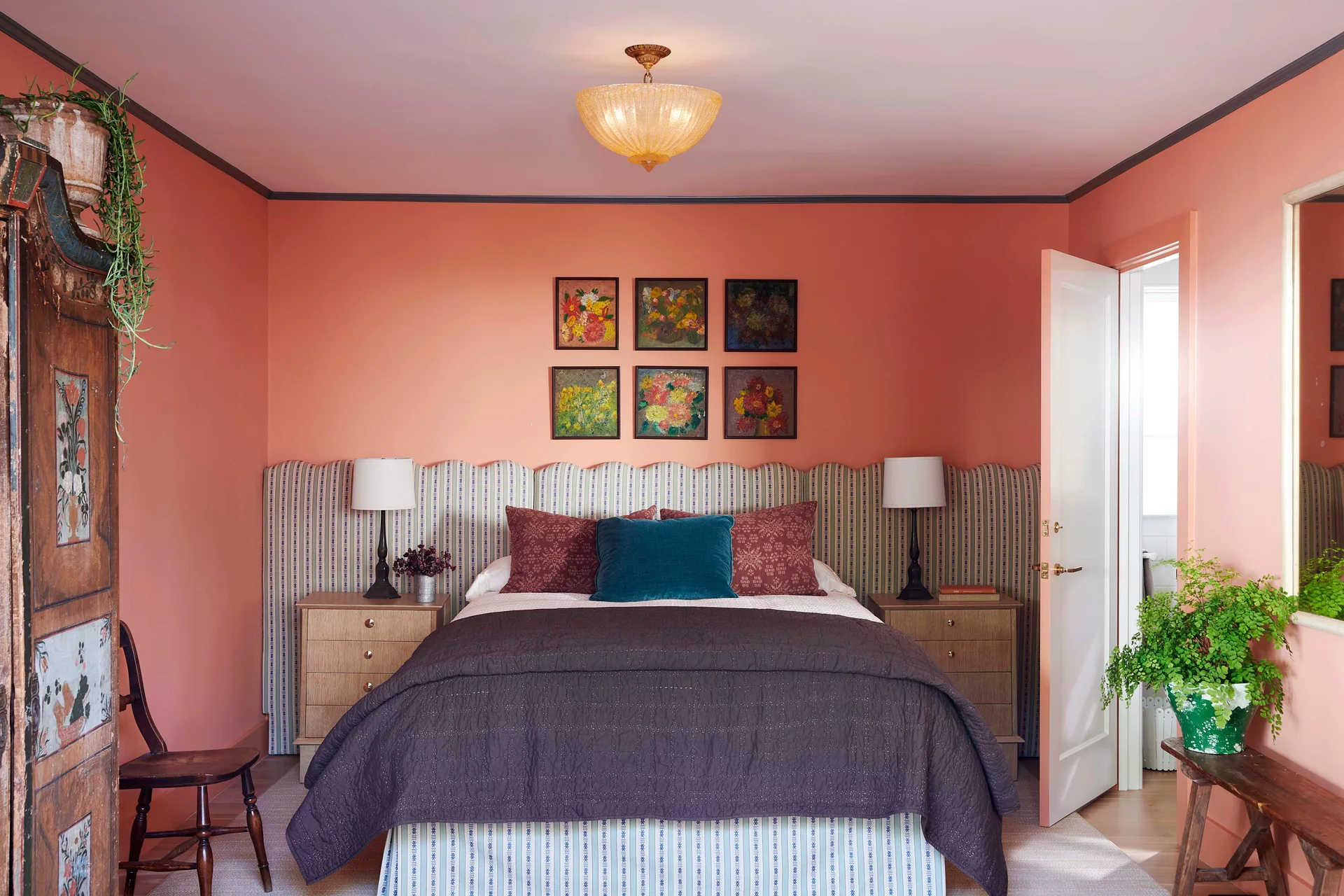 A bedroom features coral walls, an extra-wide striped headboard behind the bed, wooden nightstands with lamps, and a grid of six framed floral prints.